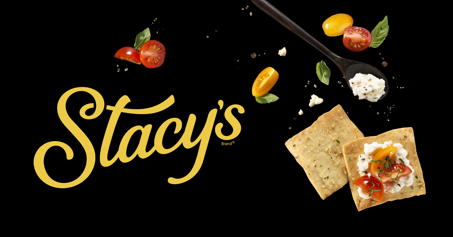 Stacy's logo with scattered cherry tomatoes, basil leaves, crackers topped with cheese and tomatoes, and a spoonful of cheese on a black background.
