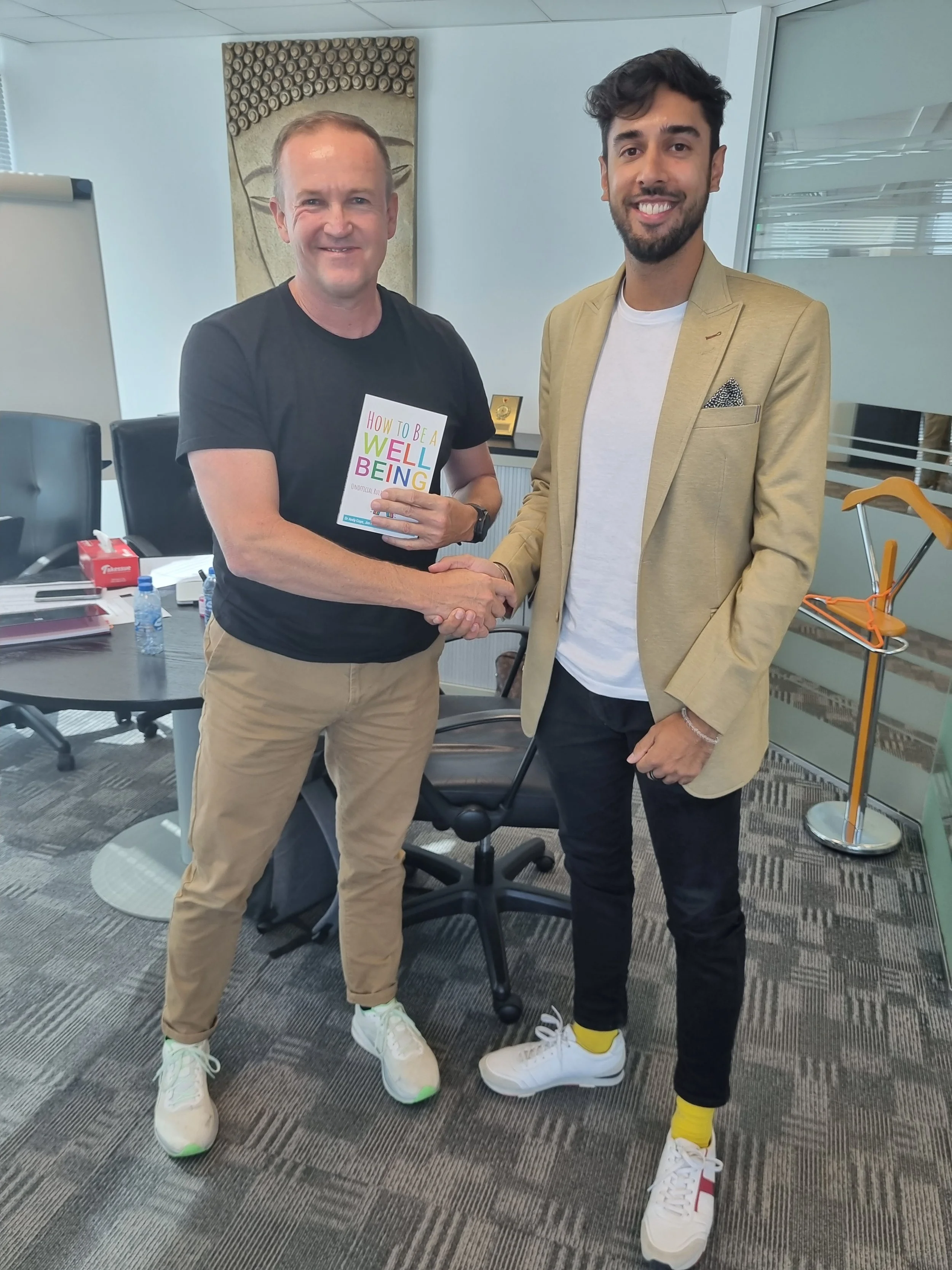 Two men shaking hands in an office space, both wearing casual shoes with bright yellow accents. One man is holding a book titled 'How to Be a Well Being'. The office has a gray carpet, chairs, and a desk with items including a water bottle, tissue box, and papers.