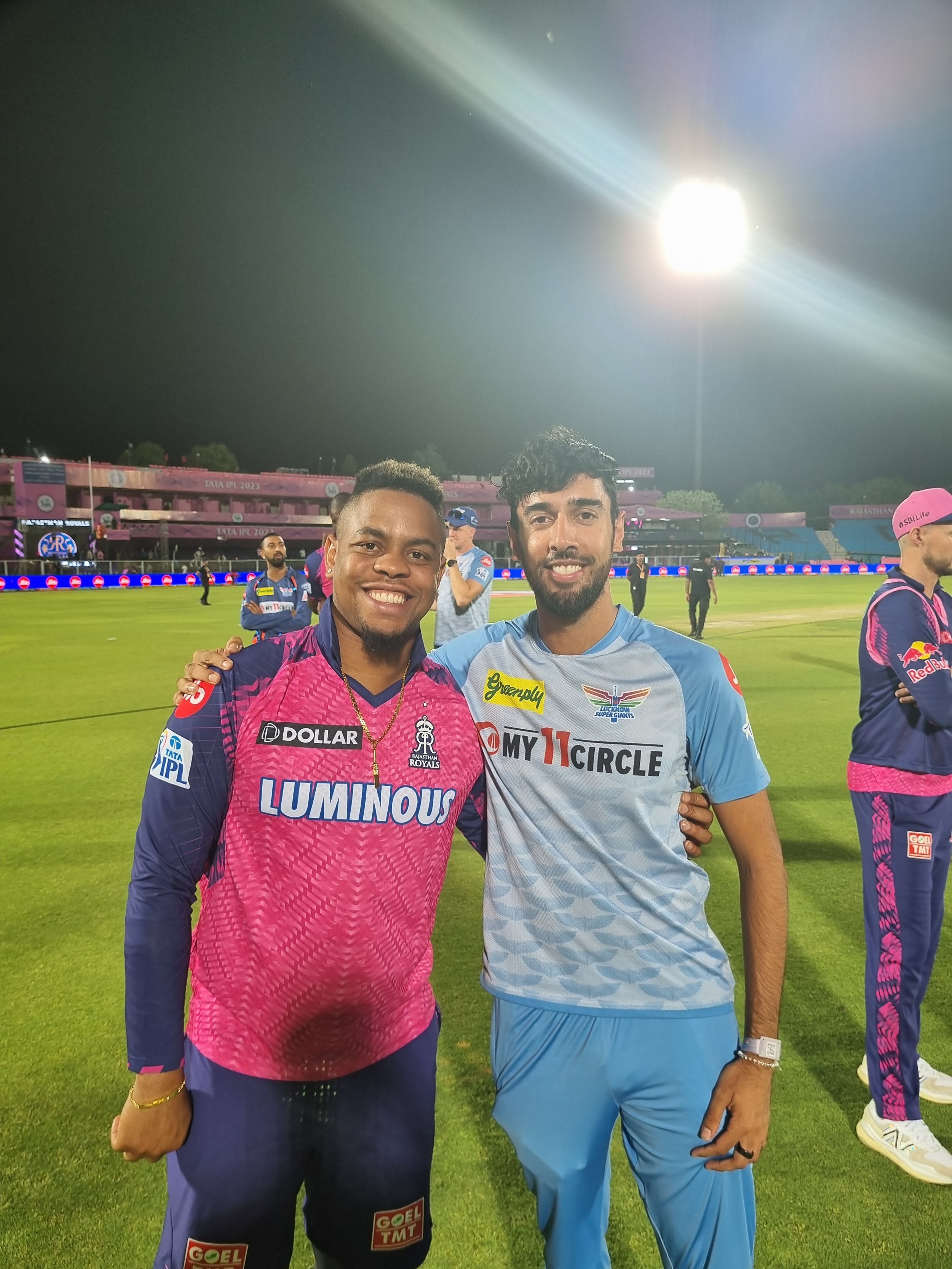 Two male cricket players standing on a cricket field at night, smiling and posing for a photo after a match, with other players in the background.