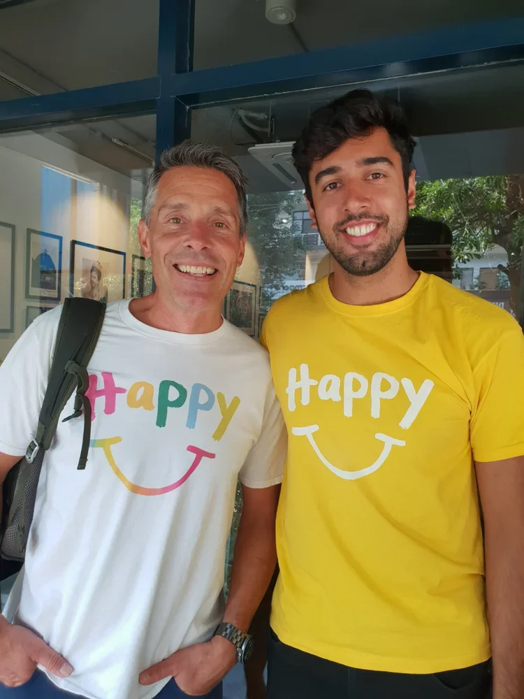 Two men smiling, one older with gray hair and the other younger with dark hair, standing indoors. Both are wearing white and yellow T-shirts with the word "Happy" and a smiley face printed on them.