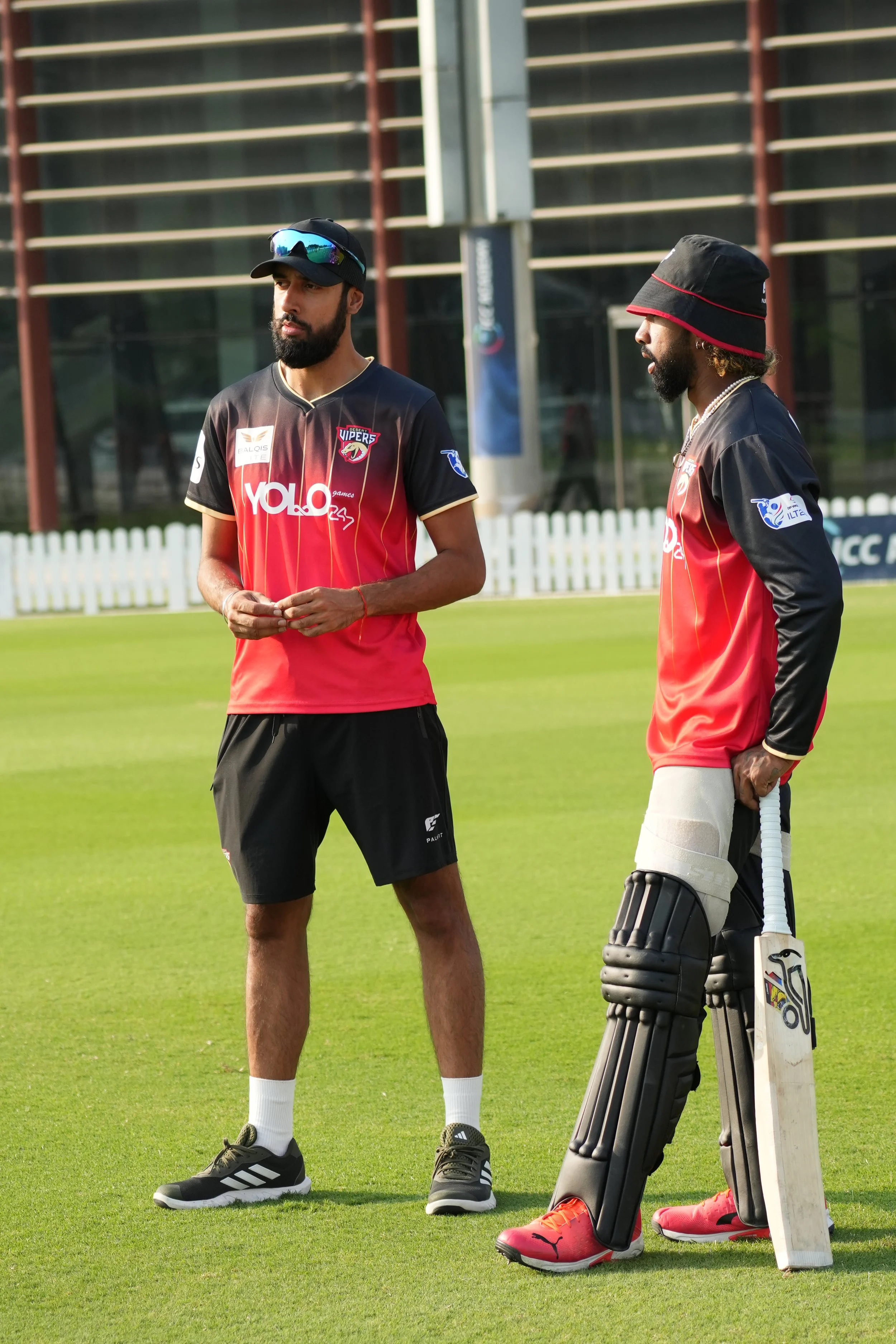 Two cricket players on a field, one in batting gear and the other in casual sportswear, standing and talking.
