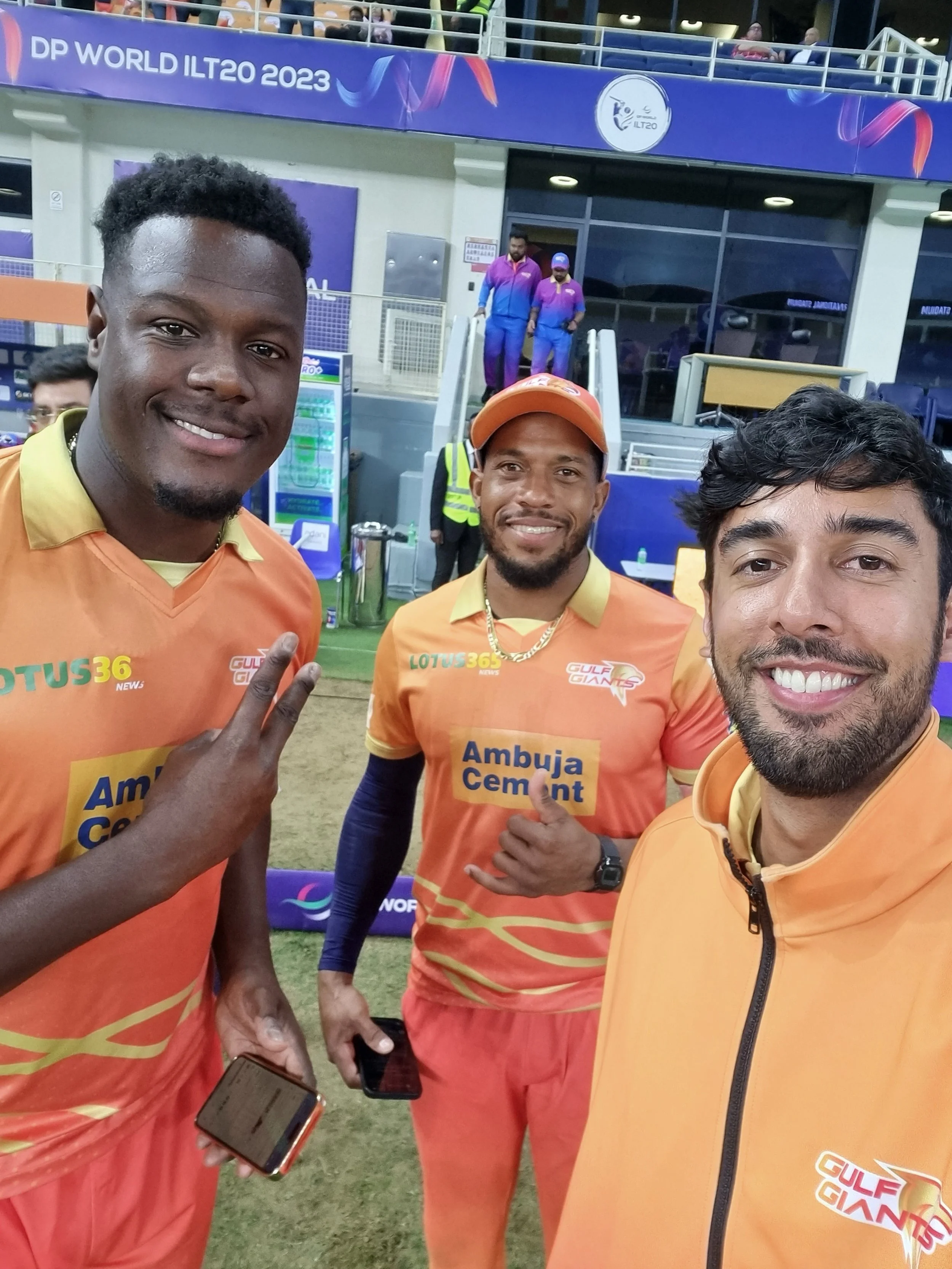 Three men in orange uniforms with sponsor logos taking a selfie at a cricket stadium. Two of them are making peace signs, and one is giving a thumbs-up. Behind them, there are spectators and officials in purple and blue uniforms, with the sign 'DP World ILT20 2023' visible.