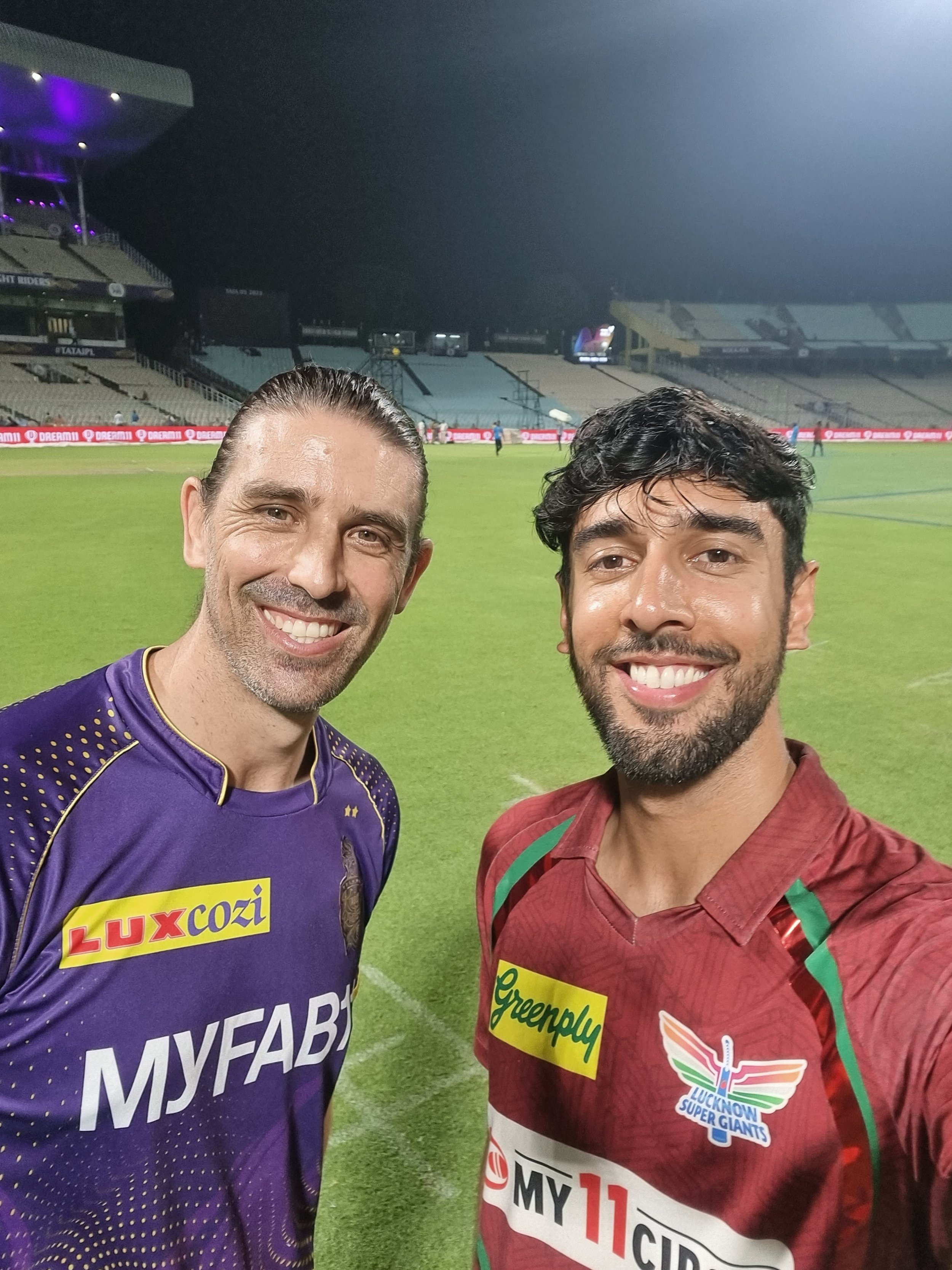 Two smiling men taking a selfie at a cricket stadium after a match. One man is wearing a purple cricket jersey with yellow and white accents, and the other is wearing a maroon jersey with green and yellow accents and a logo that says "Lucknow Super Giants" and "Greenply".