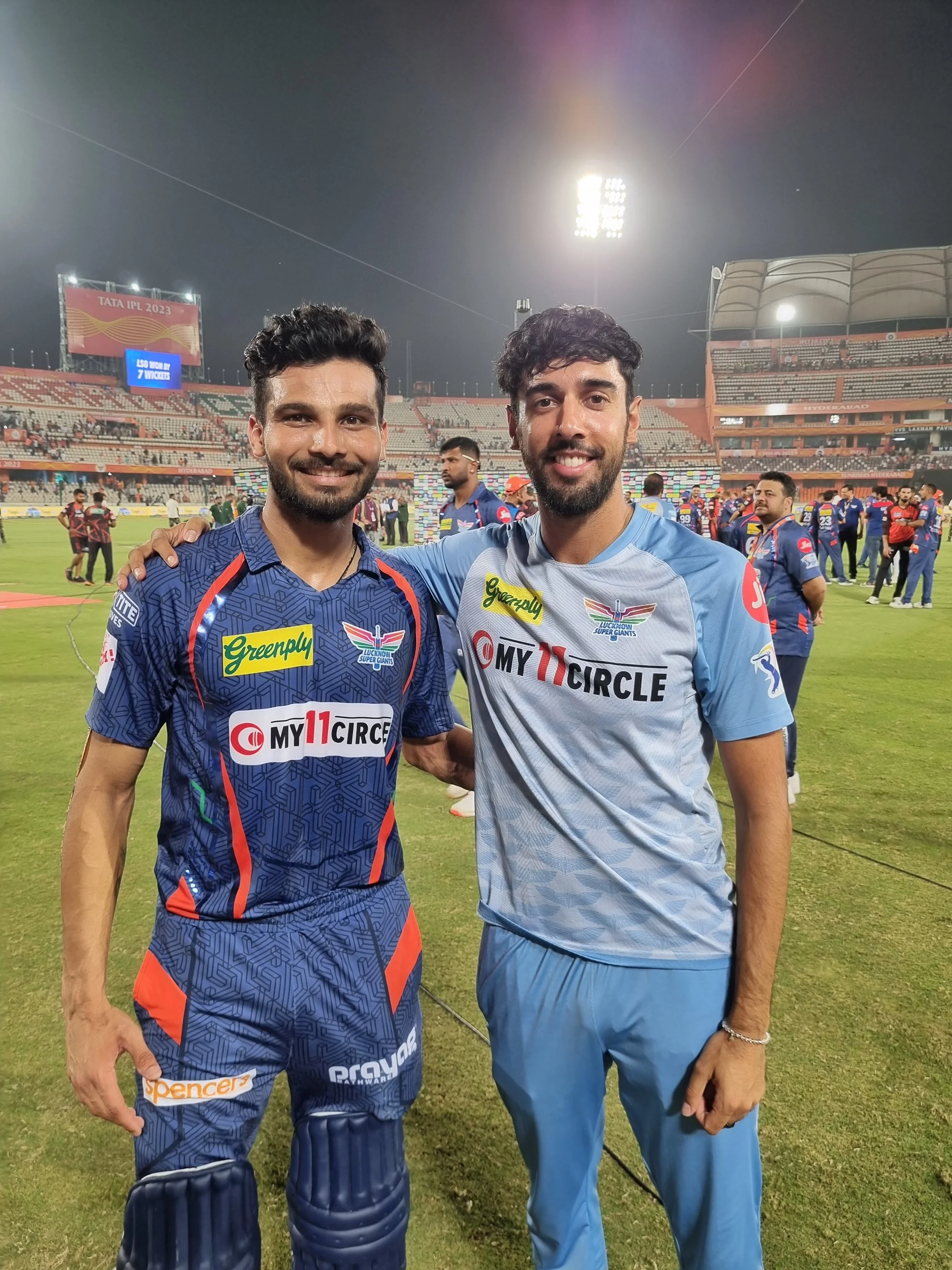Two male cricket players smiling and posing for a photo on a cricket field after a match, with one wearing a blue and orange uniform and the other in a light blue uniform, in a stadium at night.