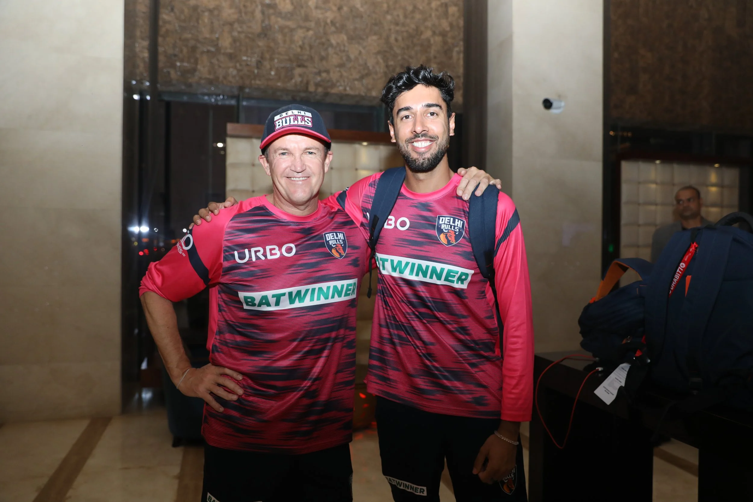 Two men dressed in pink sports jerseys with logos, smiling and posing for a photo indoors, one has his hand on the other's shoulder, in a hotel or conference setting.