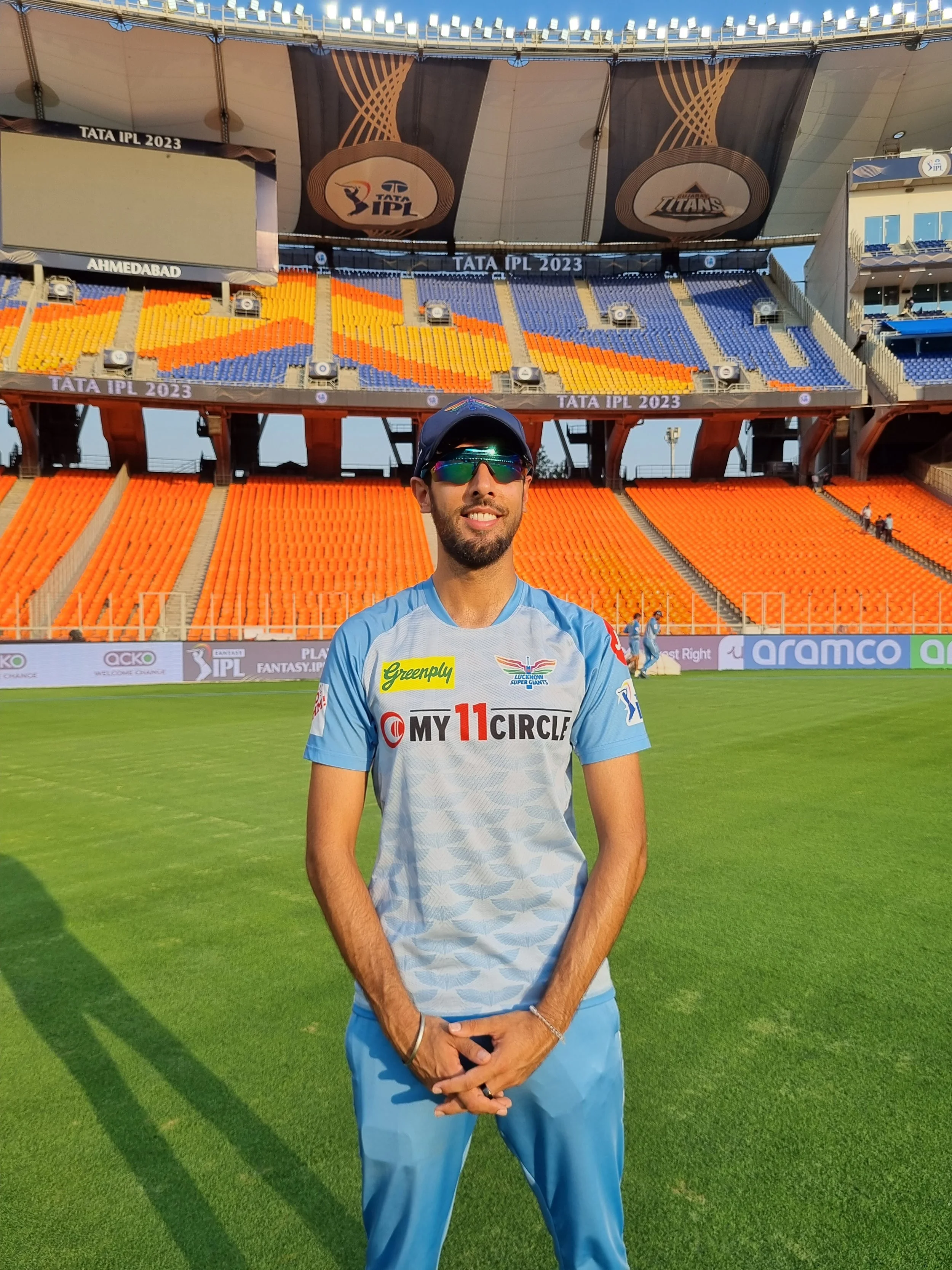 A man wearing sunglasses and a cricket uniform standing on a cricket field with empty stadium seats and banners in the background.