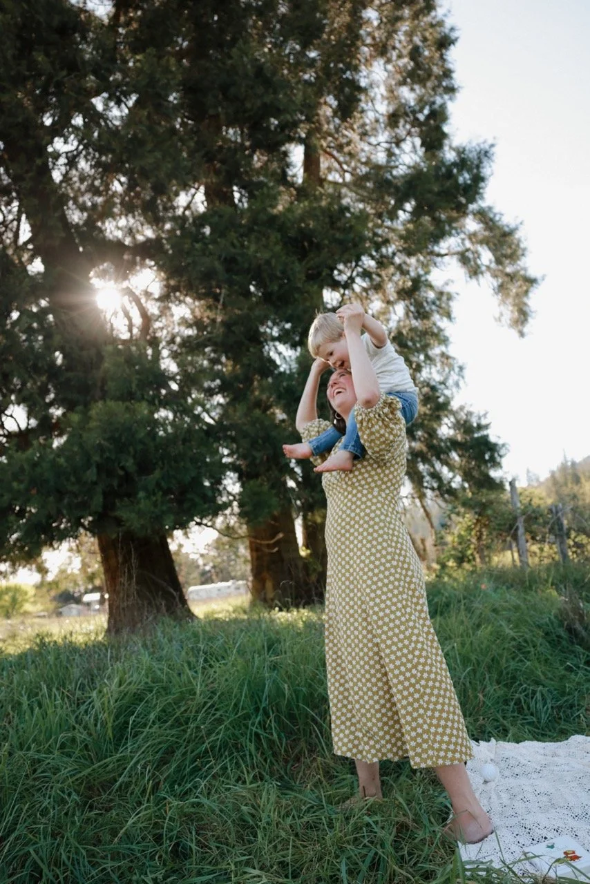 A woman lifting a young boy on her shoulders outdoors in a grassy area with tall trees and sunlight filtering through the branches.