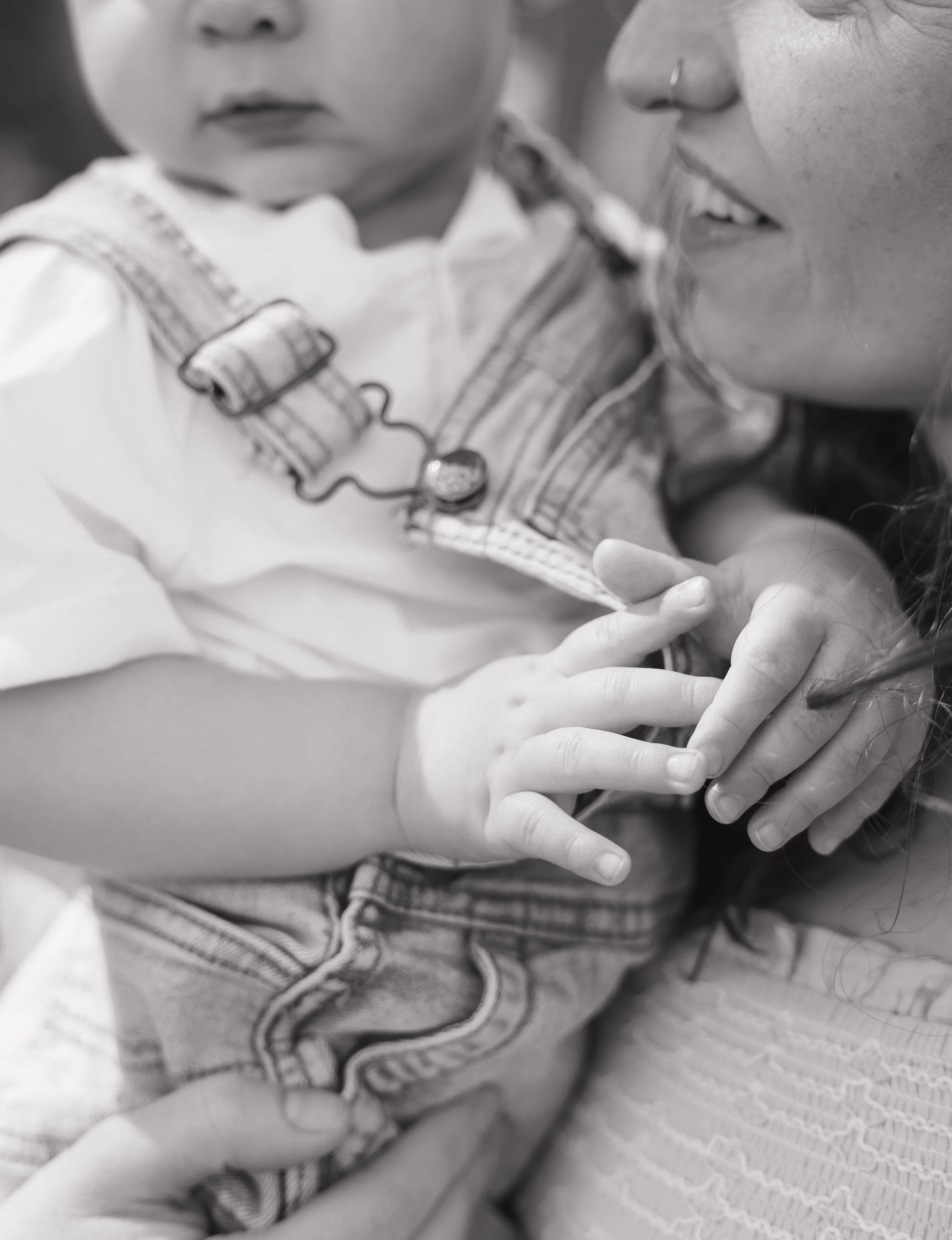 A close-up black-and-white photo of a young child with curly hair and another person holding the child's hands and a lollipop near their mouth.