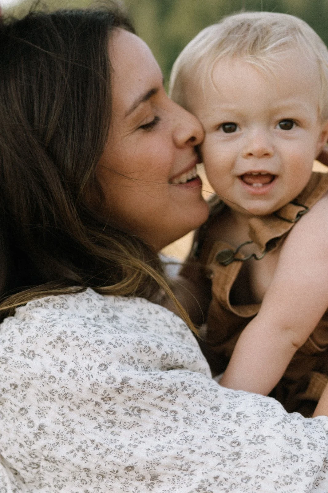 A woman holding a young blonde-haired boy close, smiling and looking at the camera, with her face near his. The boy is wearing a brown sleeveless shirt and has a curious expression.