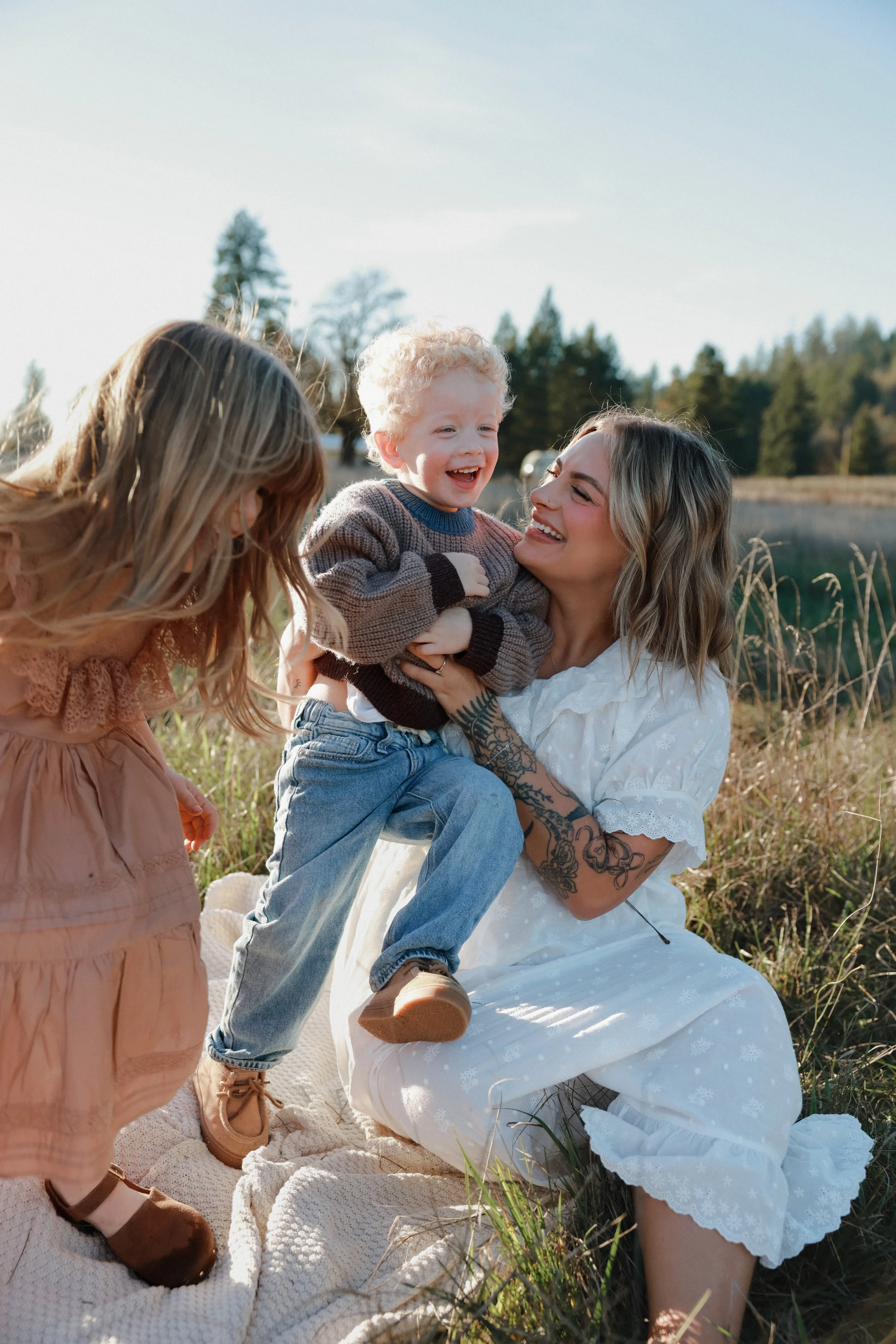 A woman smiles while holding a young boy, who is laughing, outdoors near a lake and trees, with a girl in a pink dress and brown shoes nearby.