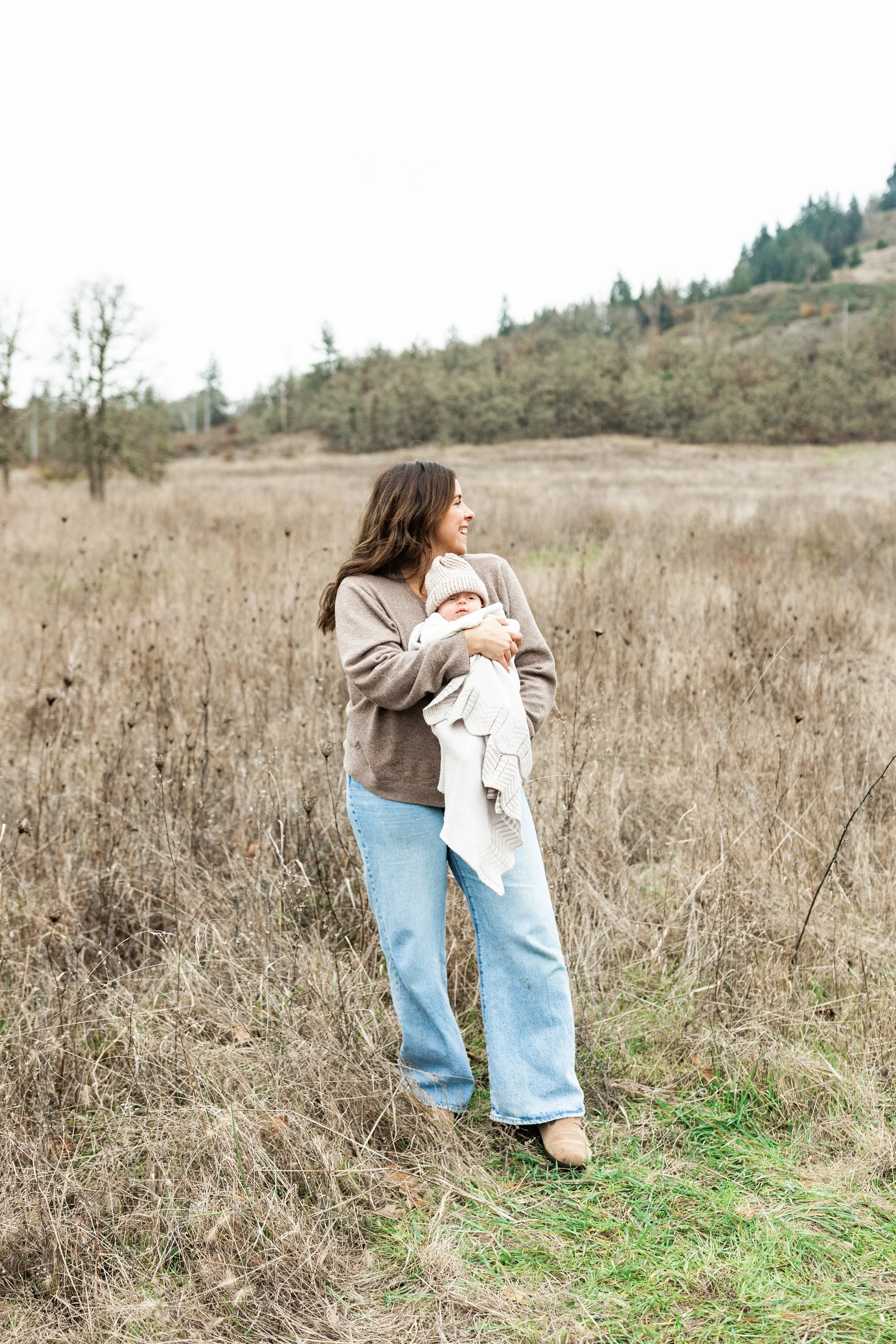 A woman holding a baby in a grassy field with hills and trees in the background.