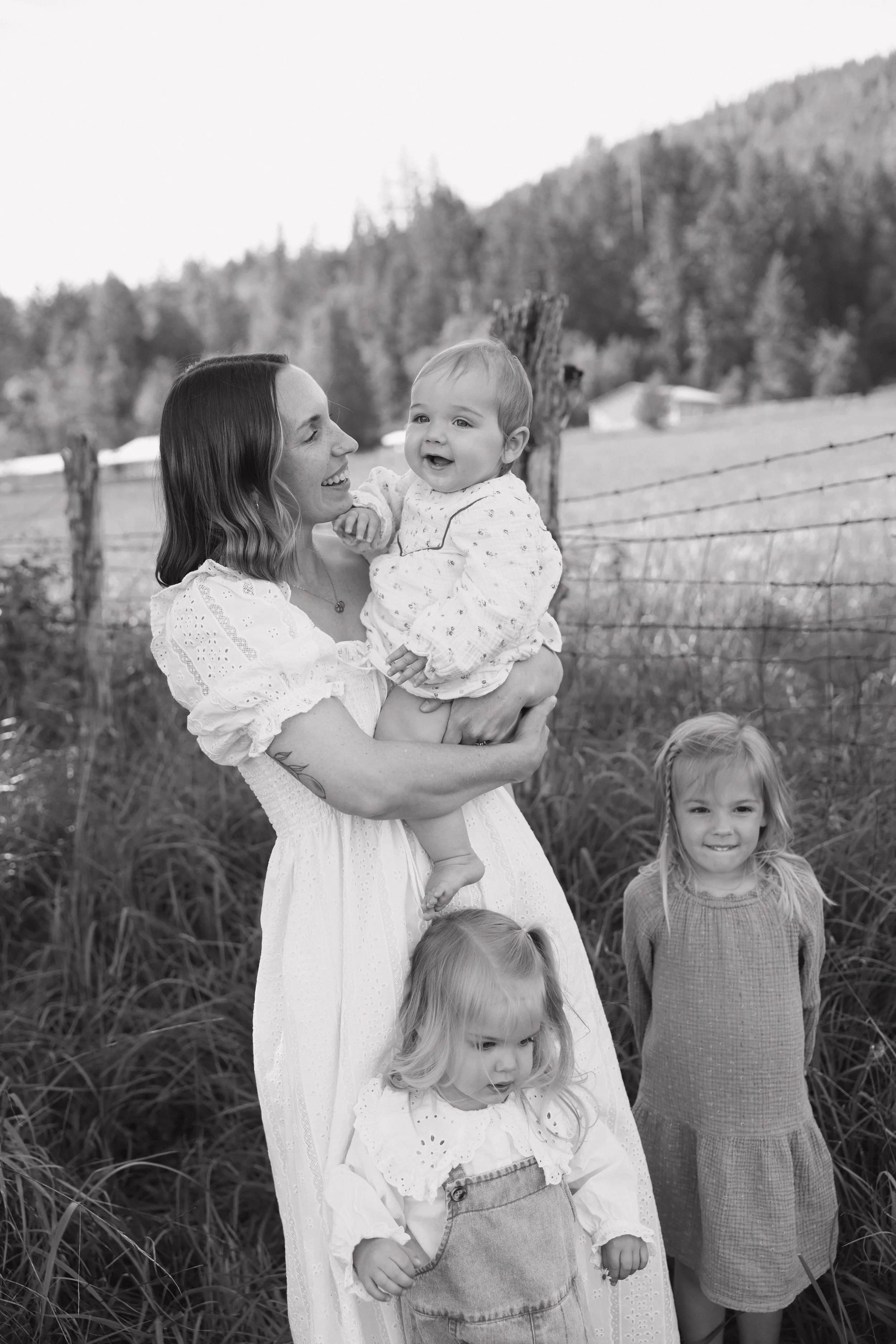 A woman holding a smiling baby girl in a rural outdoor setting with a fence, trees, and mountains in the background. Other children are standing and sitting nearby.