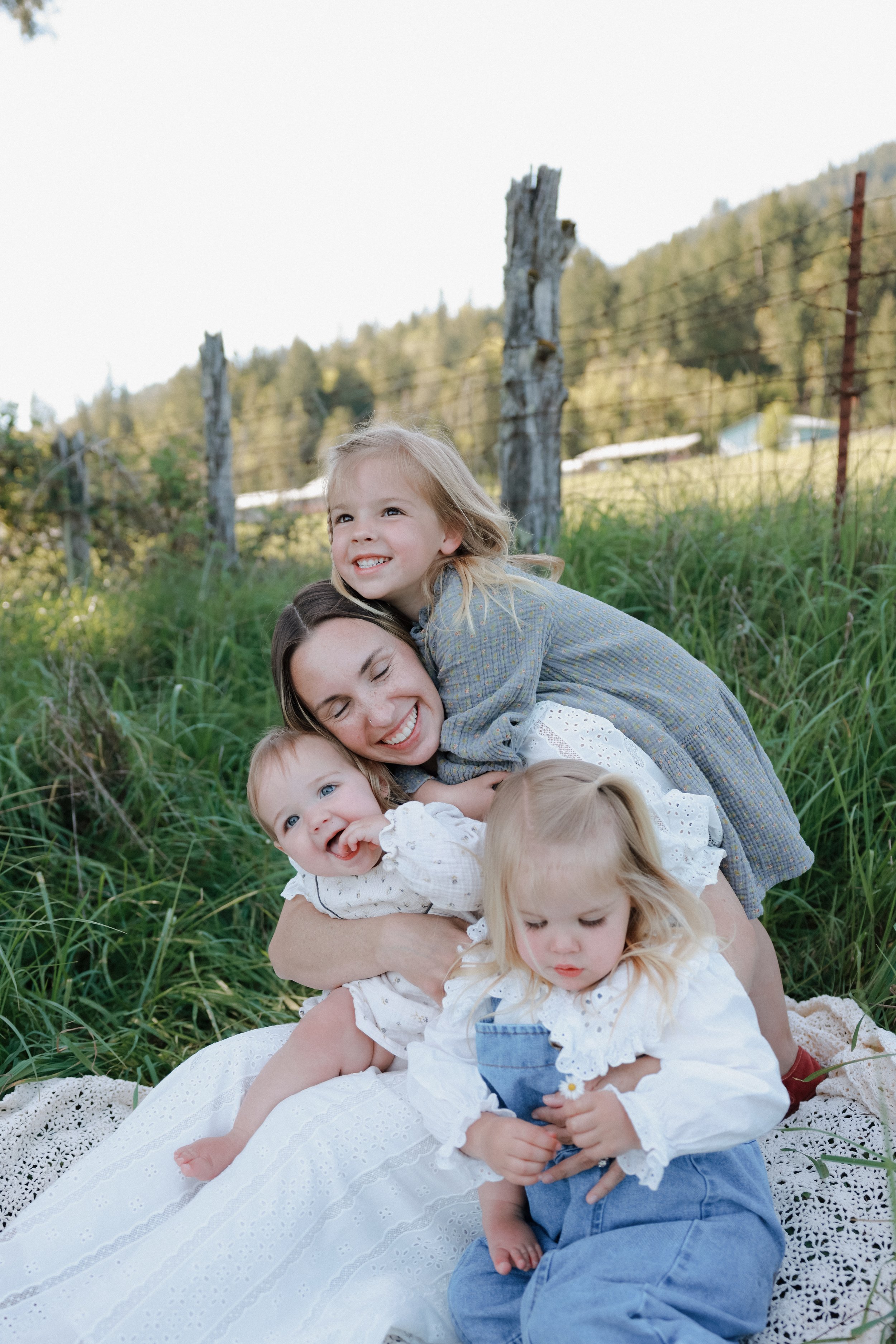 A woman with brown hair and five young children with blonde hair lying on a white lace blanket in a grassy outdoor setting. One girl is on top of the woman, smiling at the camera. Other children are playing or resting, with a rustic wooden fence and 