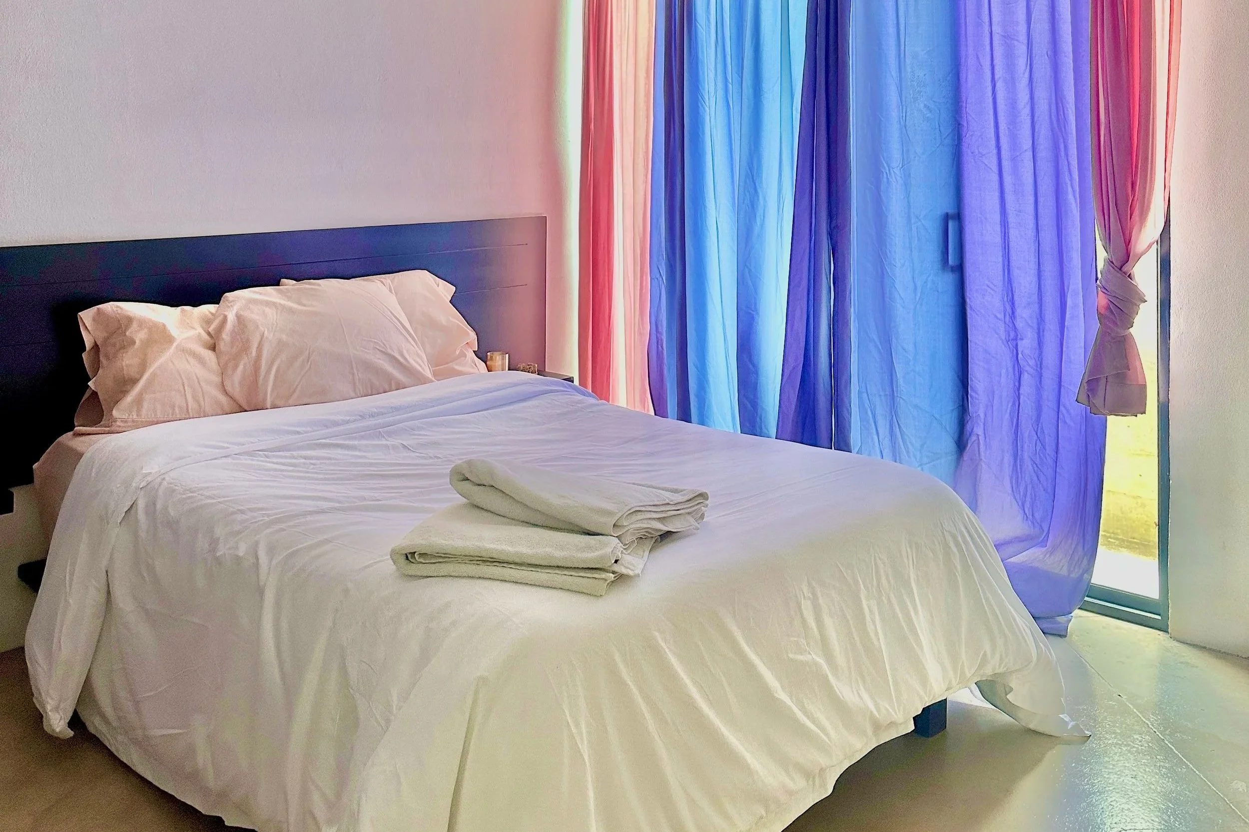 A neatly made bed with beige pillows and white sheets, placed in front of colorful curtains in shades of pink and blue, with towels folded on top of the bed.