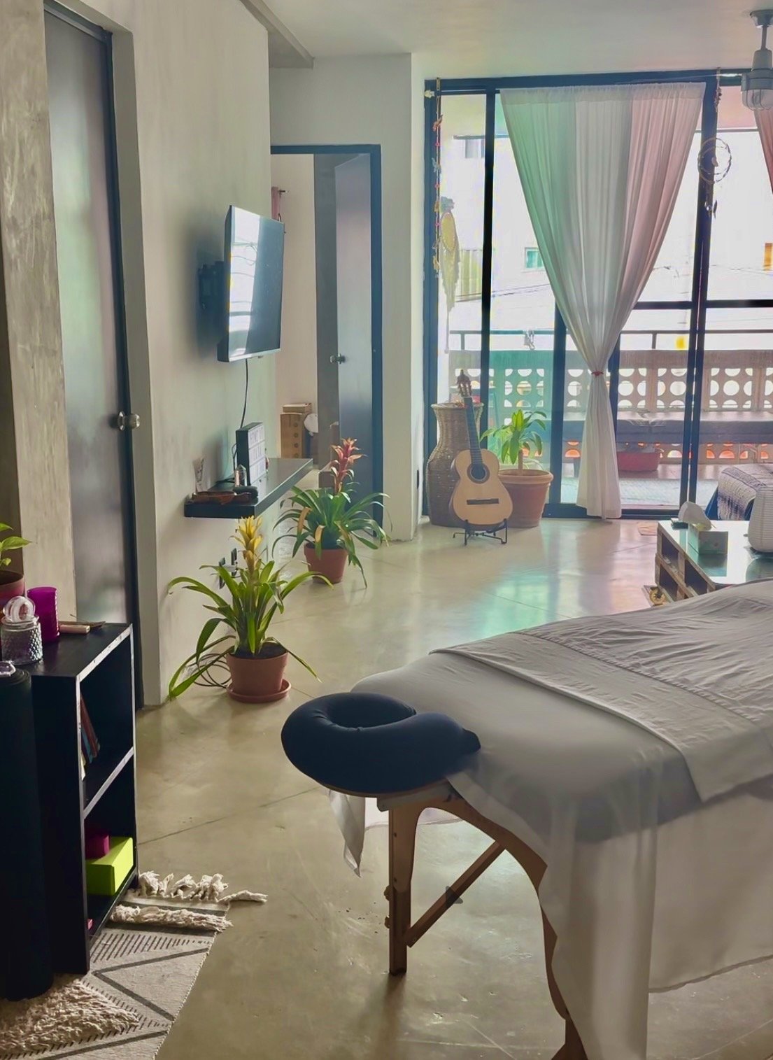 A bright, airy room with a massage or spa bed near the center, surrounded by potted plants. A large window with curtains leads to a balcony with a patterned railing. A guitar, vase, and plant are near the window. A wall-mounted TV and a small shelf with various items are on the wall. The room has a modern, relaxed decor with natural light streaming in.