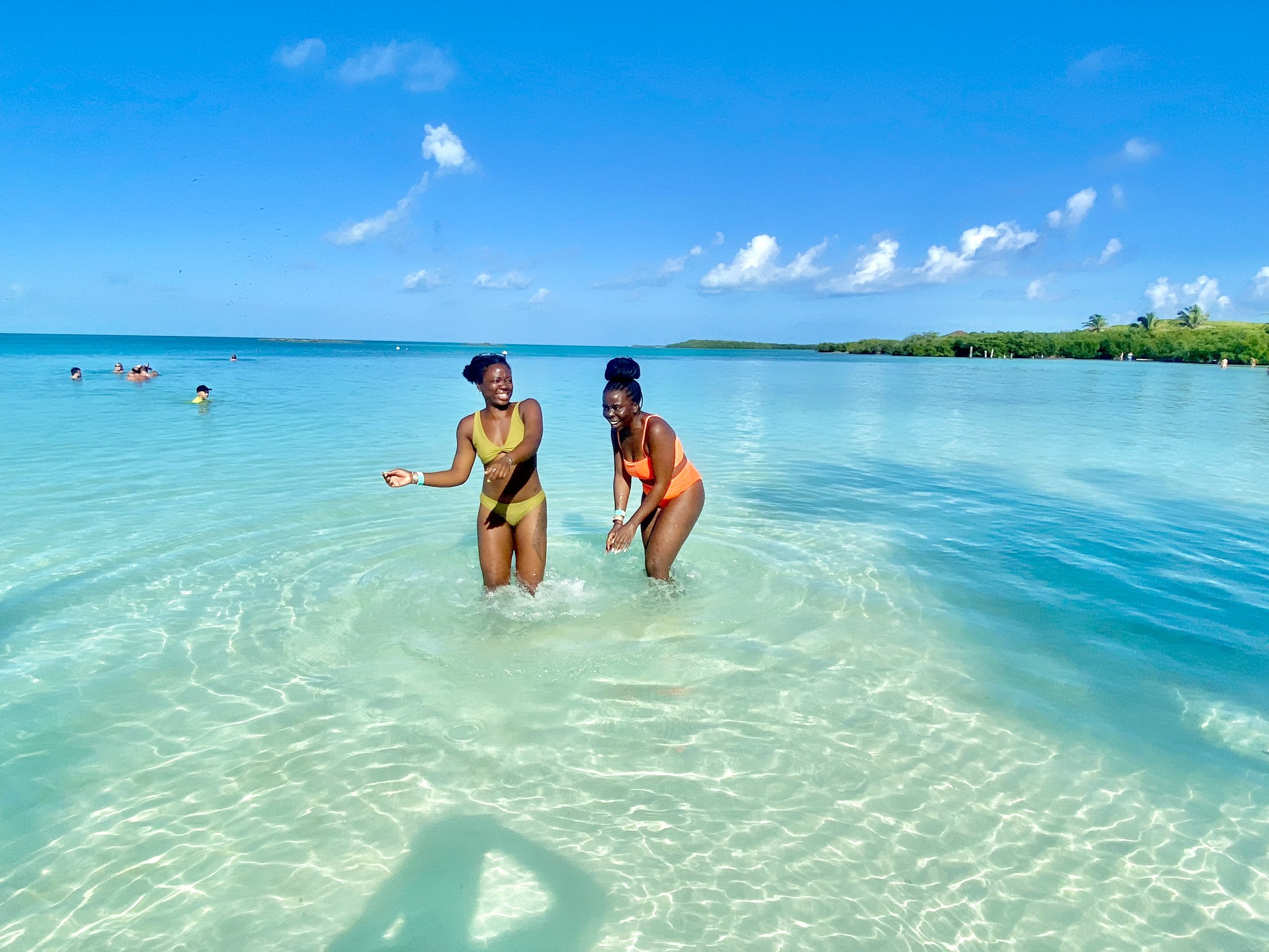 Two women in swimsuits enjoying time in shallow turquoise water on a sunny beach with a clear blue sky and distant land.