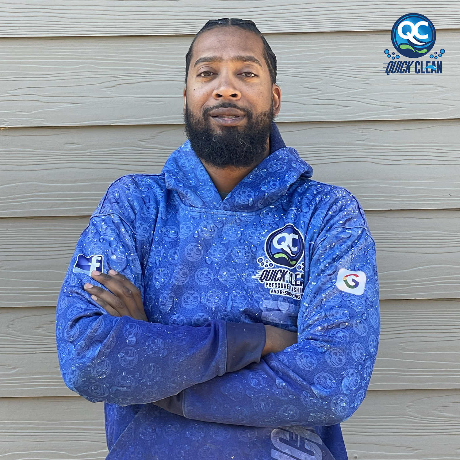 A man with a beard and braids wearing a blue logoed hoodie with the words Quick Clean. He is standing outdoors in front of a beige wooden wall with his arms crossed and looking directly at the camera.