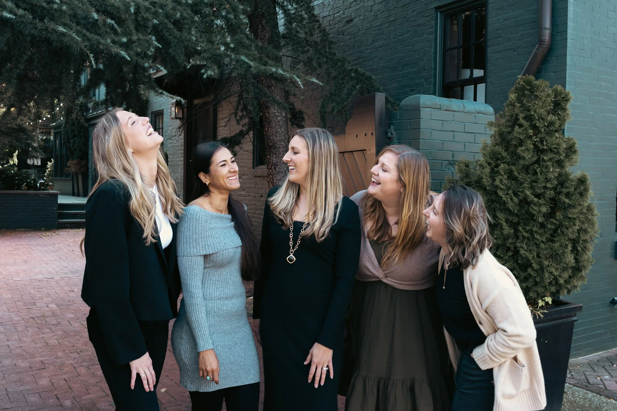 Group of six women laughing and chatting outdoors on a city sidewalk, standing near a green brick building and trees.