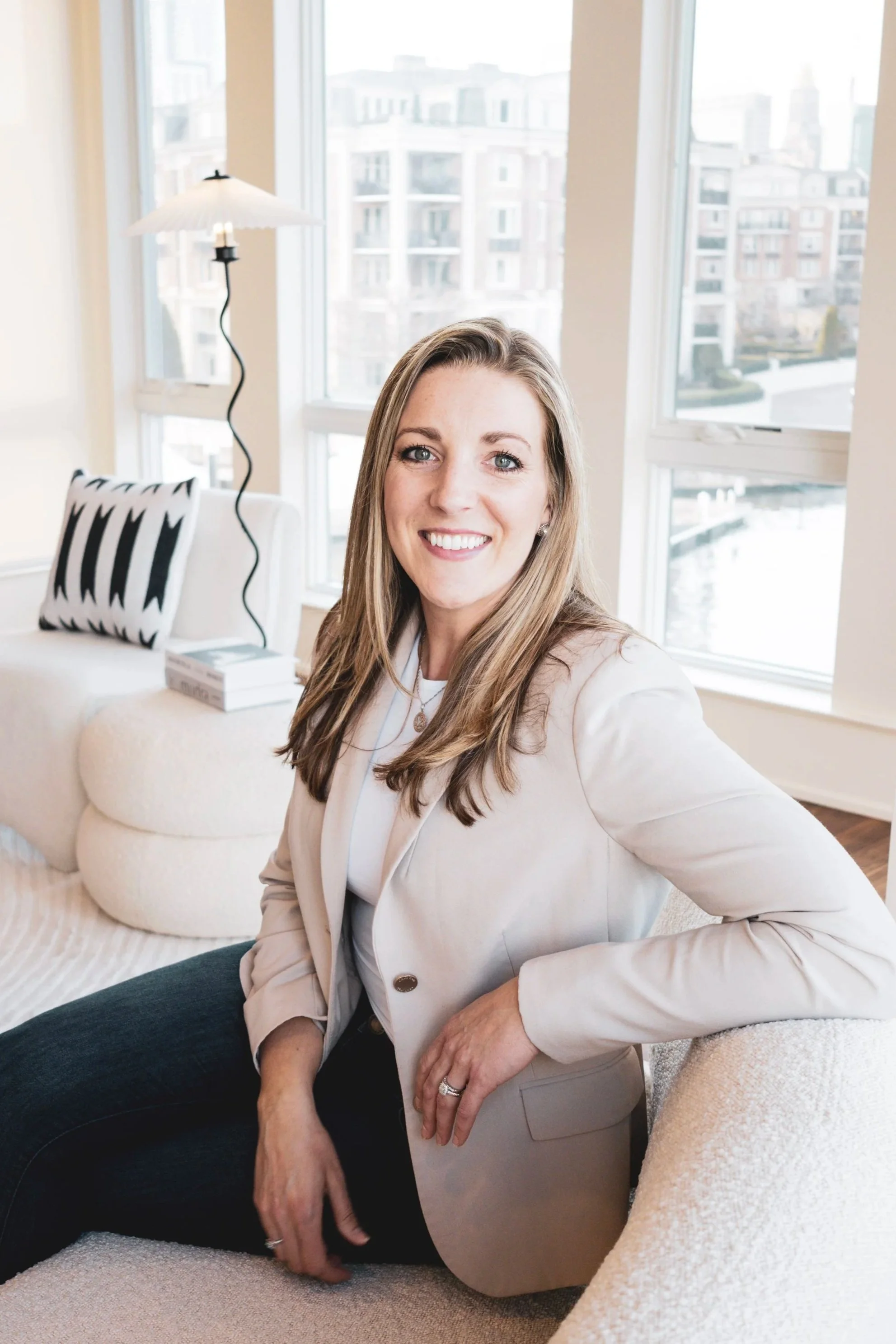 A woman with long light brown hair and blue eyes smiling while sitting on a white couch in a bright room with large windows showing city buildings.