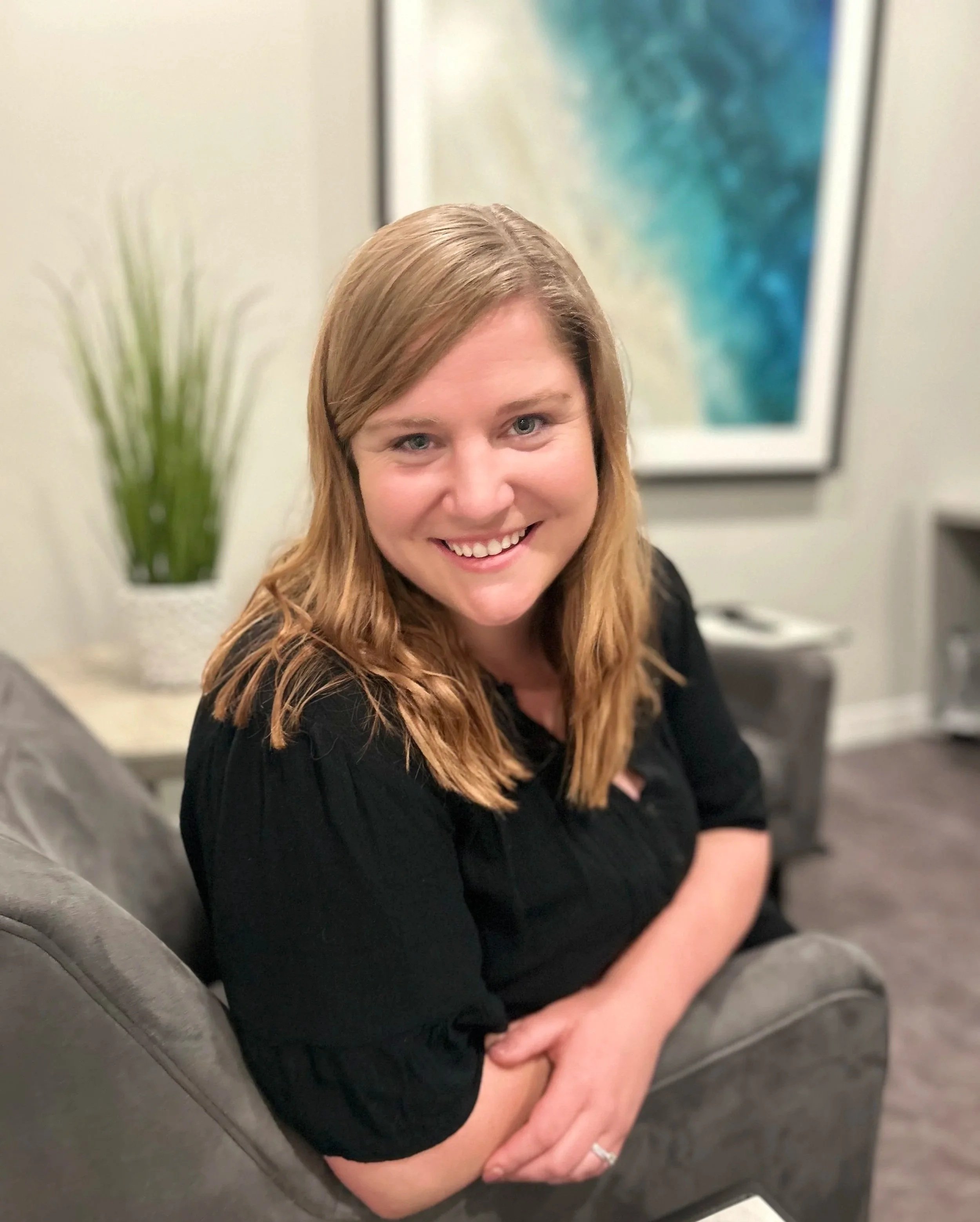 A woman with long light brown hair smiling, sitting on a gray sofa in a living room with a potted plant and abstract artwork on the wall behind her.