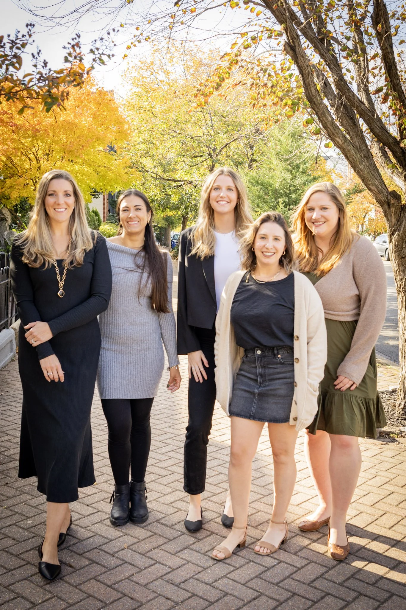 Group of five women standing outdoors on a brick sidewalk with autumn trees and sunlight in the background, smiling.