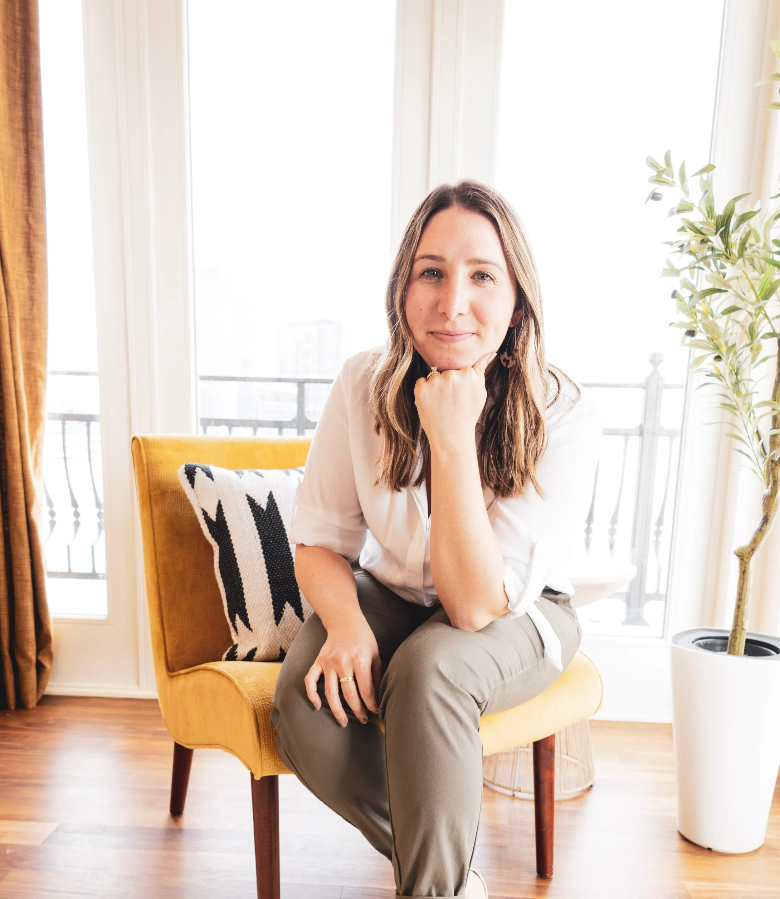 A woman with shoulder-length brown hair sitting on a yellow armchair in a bright room with large windows behind her. She is resting her chin on her hand and smiling slightly. There is a black and white patterned pillow on the chair and a potted plant to the right.