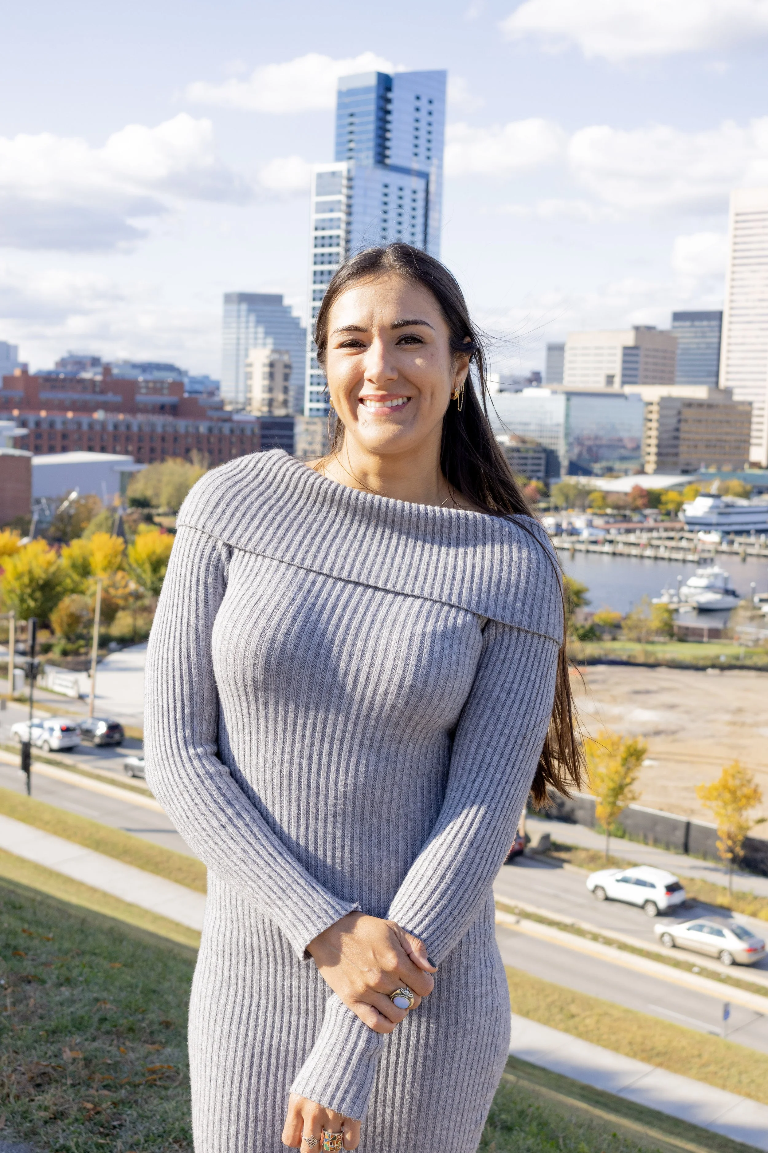A woman with long dark hair, wearing a blue shirt, smiling, with a wall and a round wall lamp in the background.