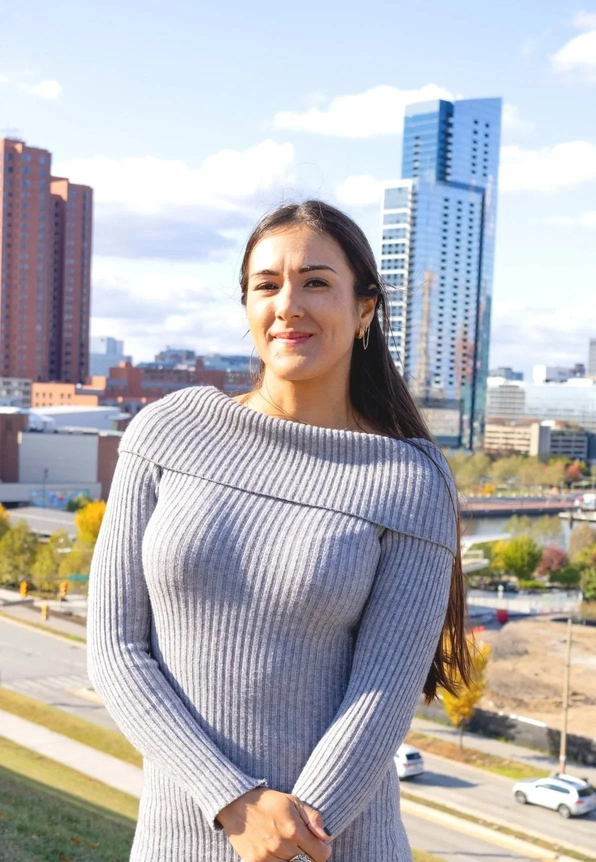 A woman with long dark hair, wearing a blue shirt, smiling, with a wall and a round wall lamp in the background.
