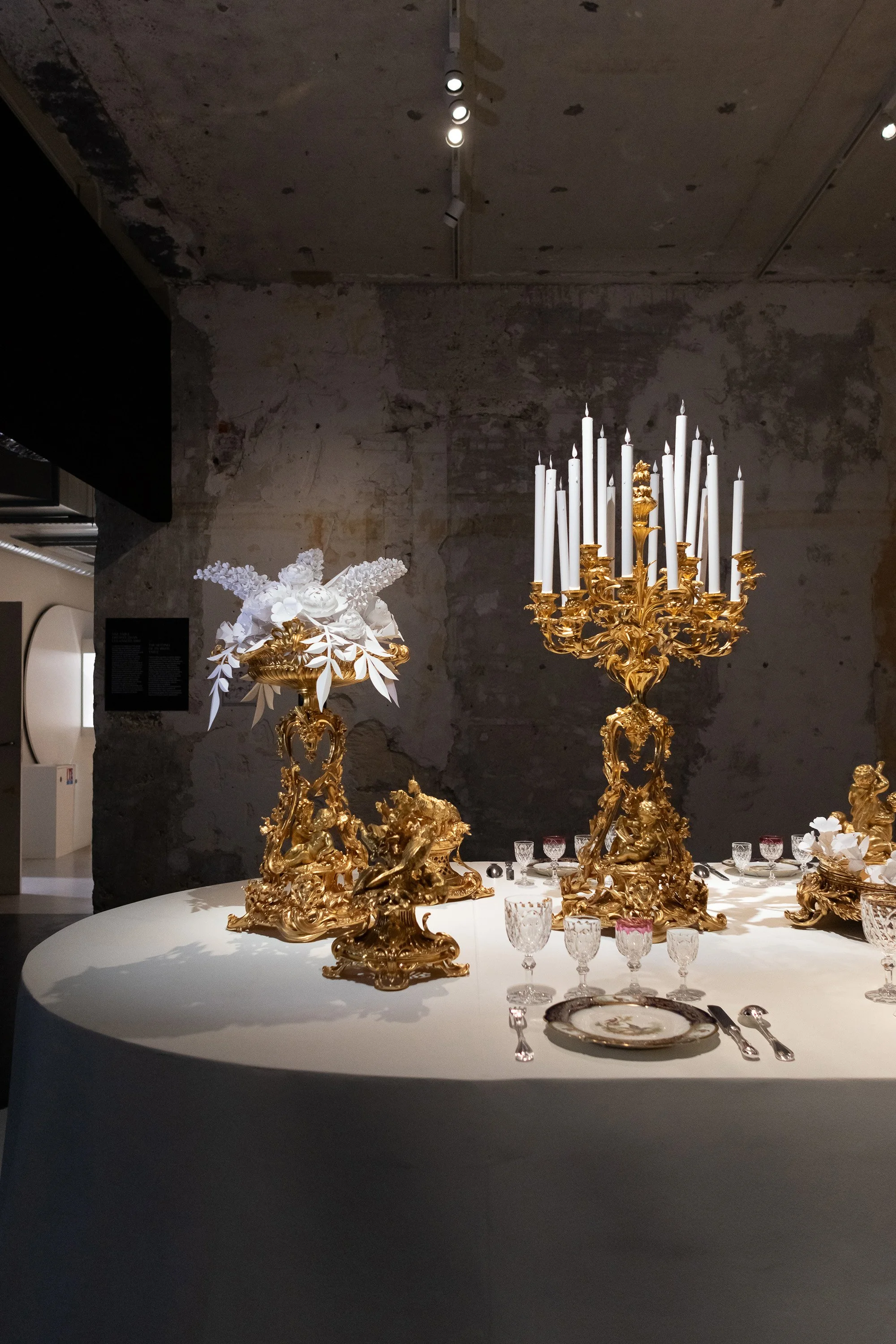 Gilded Christofle candelabra and paper floral arrangement displayed on a banquet table during “Christofle, a brilliant story” exhibition at the Musée des Arts Décoratifs, Paris.
