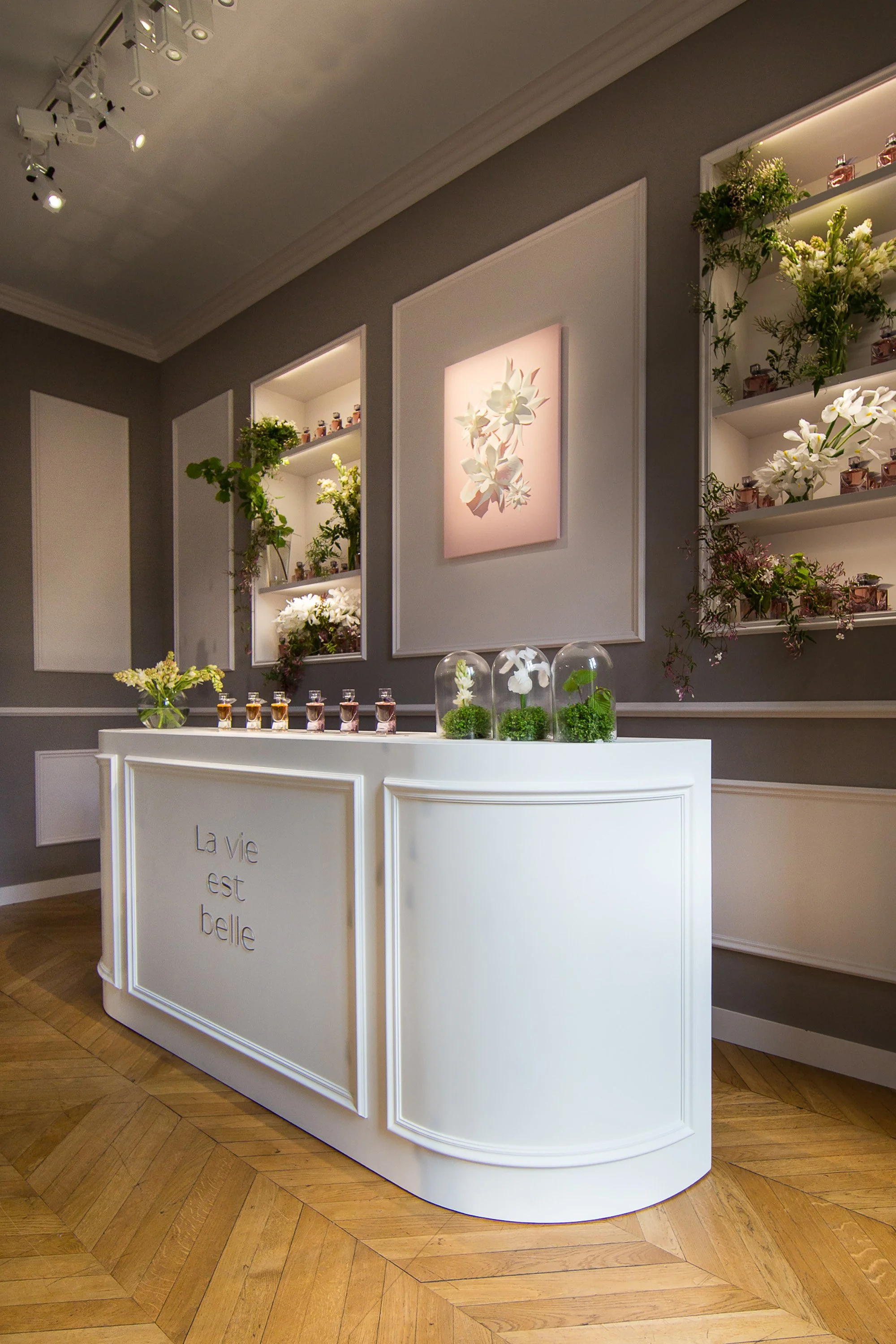 White Parisian apartment-style setting presenting La Vie Est Belle fragrances with floral arrangements and glass cloches