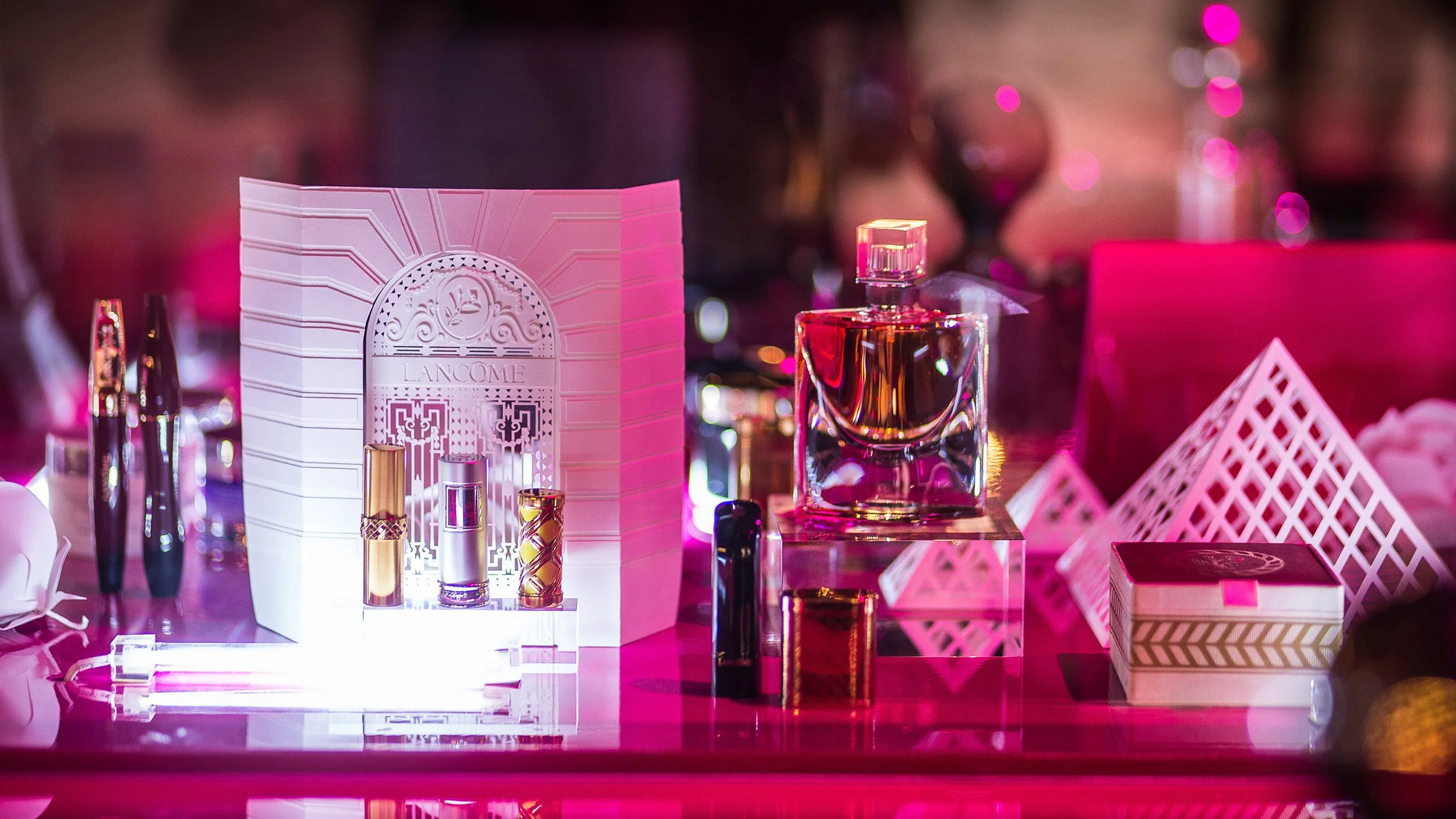 Paper Lancôme Institute façade set design with fragrance and makeup products displayed on illuminated base, pink lighting, Lancôme 80th anniversary event