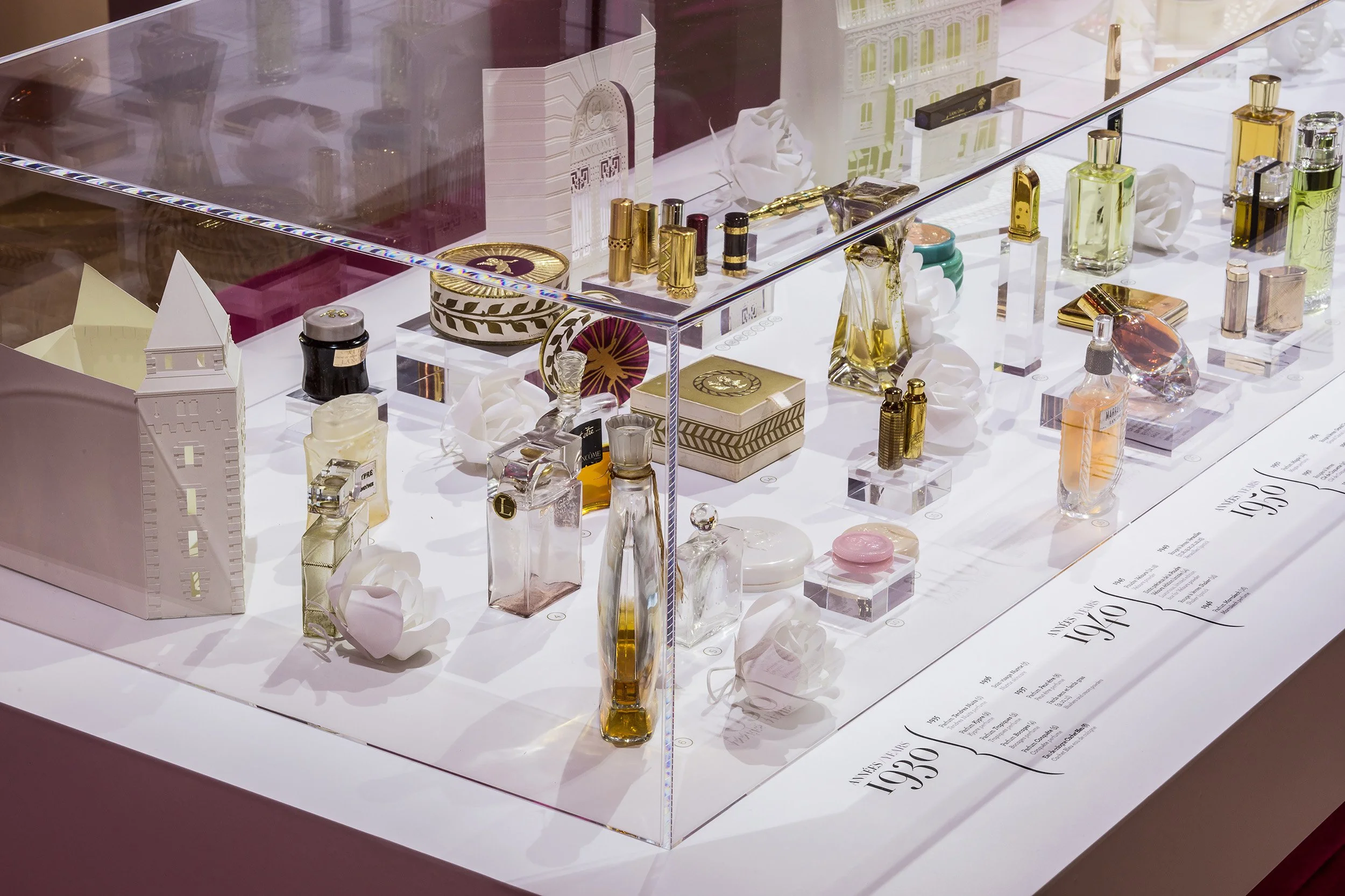 Close-up of archival Lancôme perfumes and cosmetics arranged among white paper roses and architectural elements