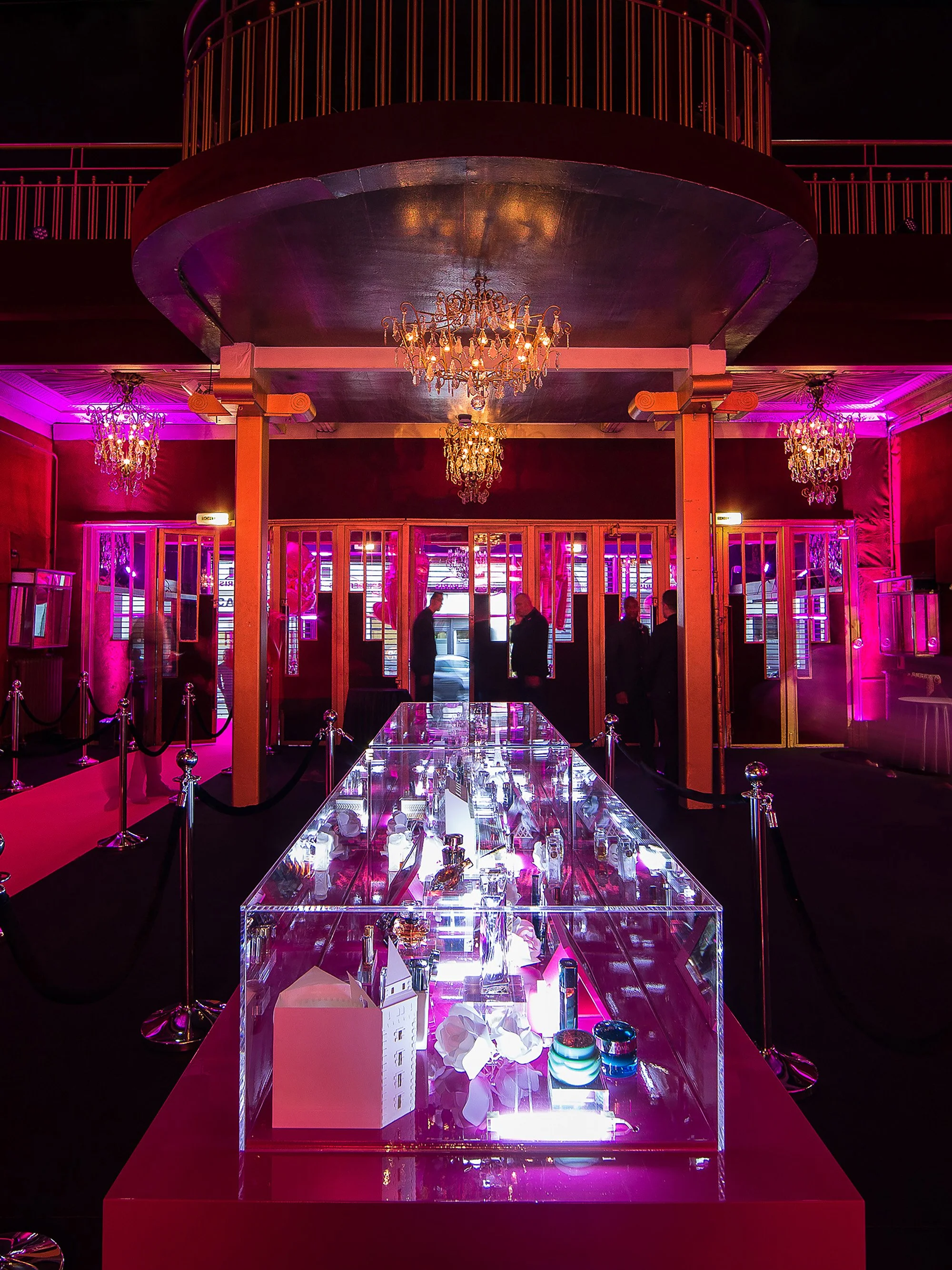 Immersive paper scenography for Lancôme’s 80th anniversary at Casino de Paris, featuring a central illuminated display case under pink lighting and chandeliers