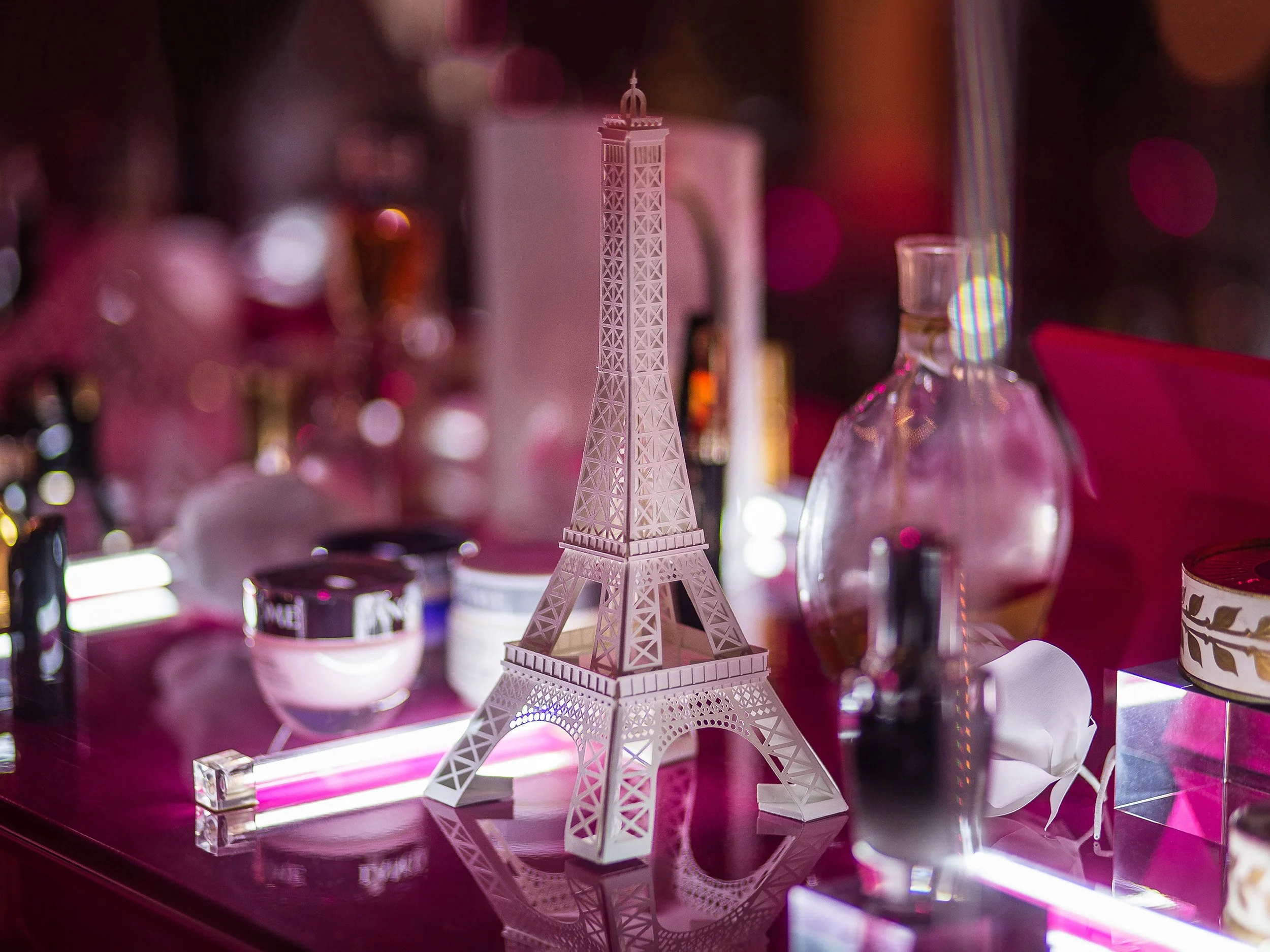 Hand-cut white paper Eiffel Tower surrounded by Lancôme fragrance and skincare products on a glossy pink display surface