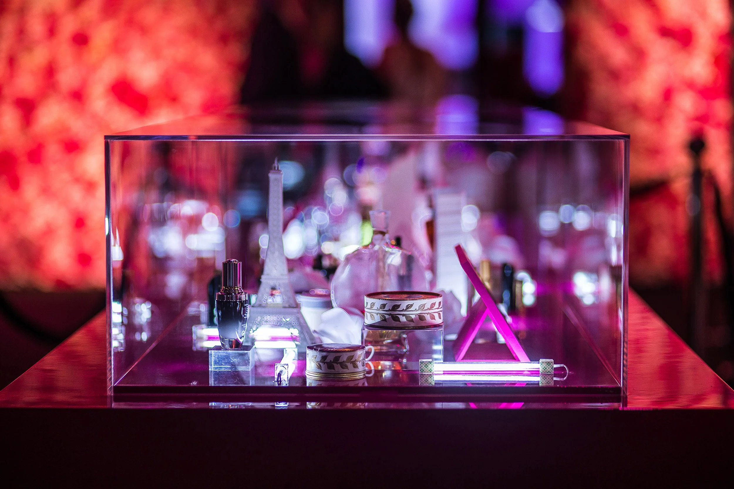 Glass display case showcasing miniature paper Parisian landmarks and Lancôme products under saturated pink event lighting
