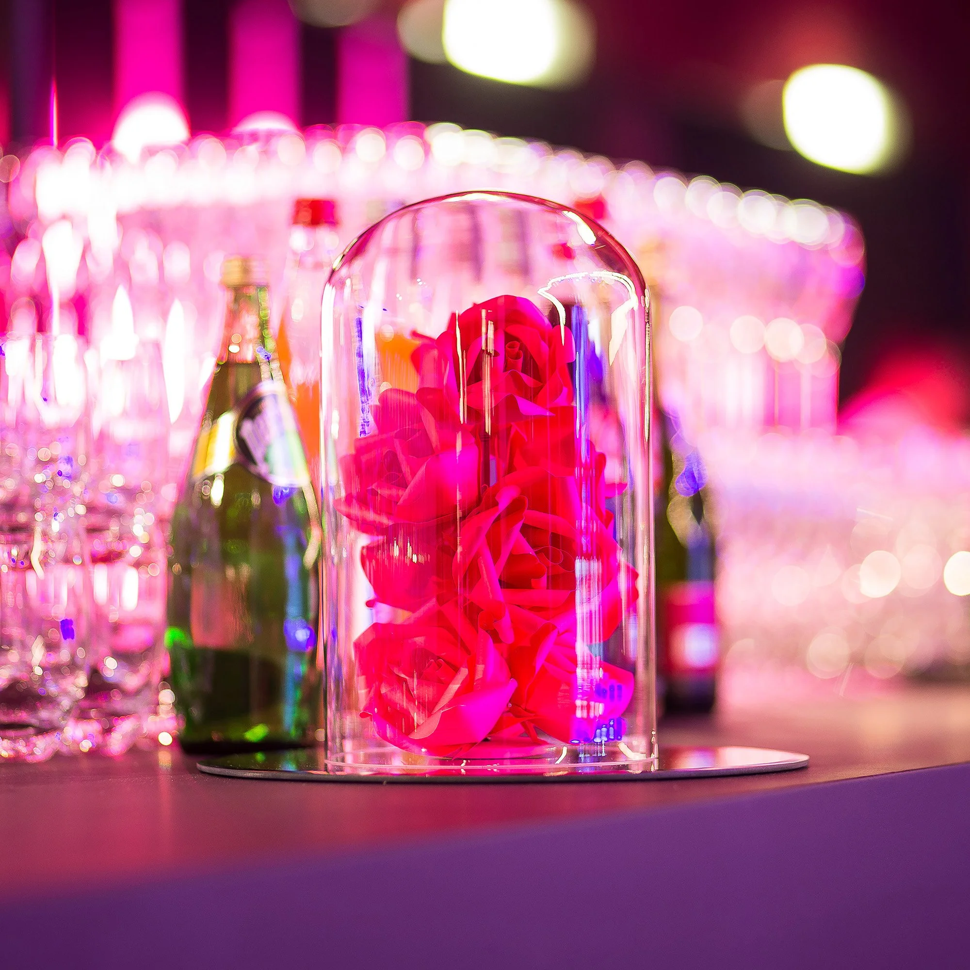 Bright pink paper roses displayed under a glass cloche on the bar, reflecting the event’s saturated lighting atmosphere