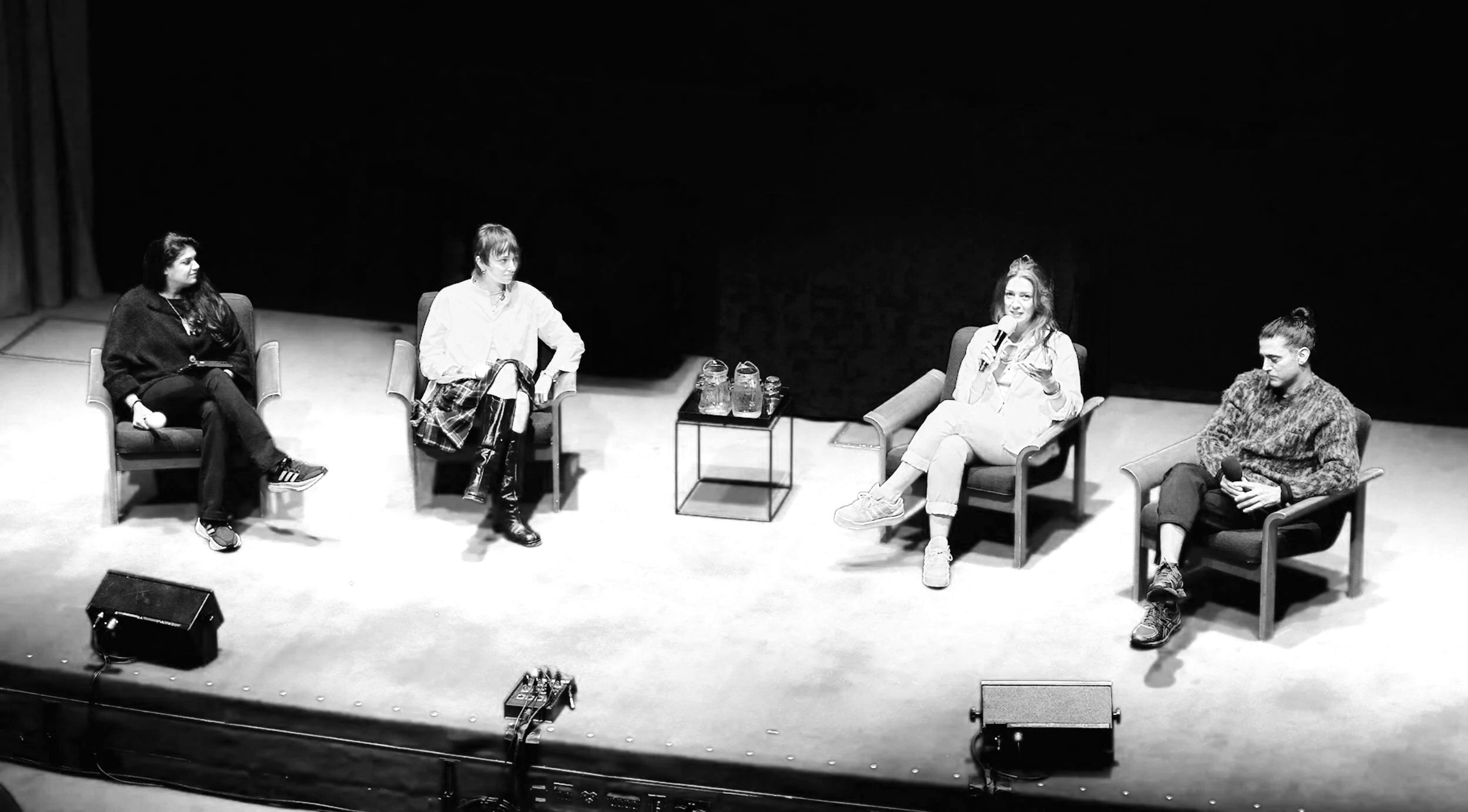Four women are seated on a stage with a red carpet, participating in a panel discussion. Three are holding microphones, and one is listening. There are water pitchers and glasses on a small table between the second and third women. Audio equipment an