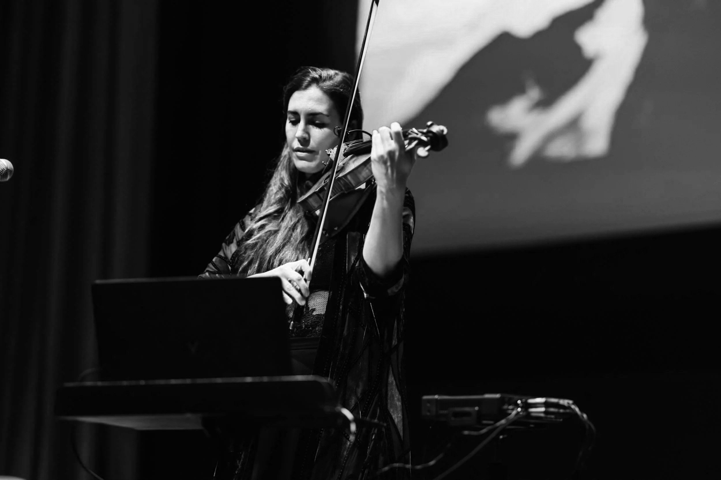 A woman playing the violin on stage with a laptop and music equipment, background featuring a large screen with a mountain landscape