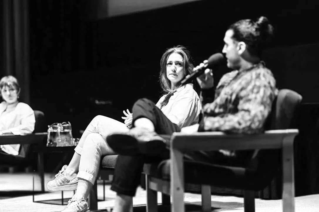 Three women participating in a panel discussion on stage, sitting in chairs with a dark background and a red carpeted floor.