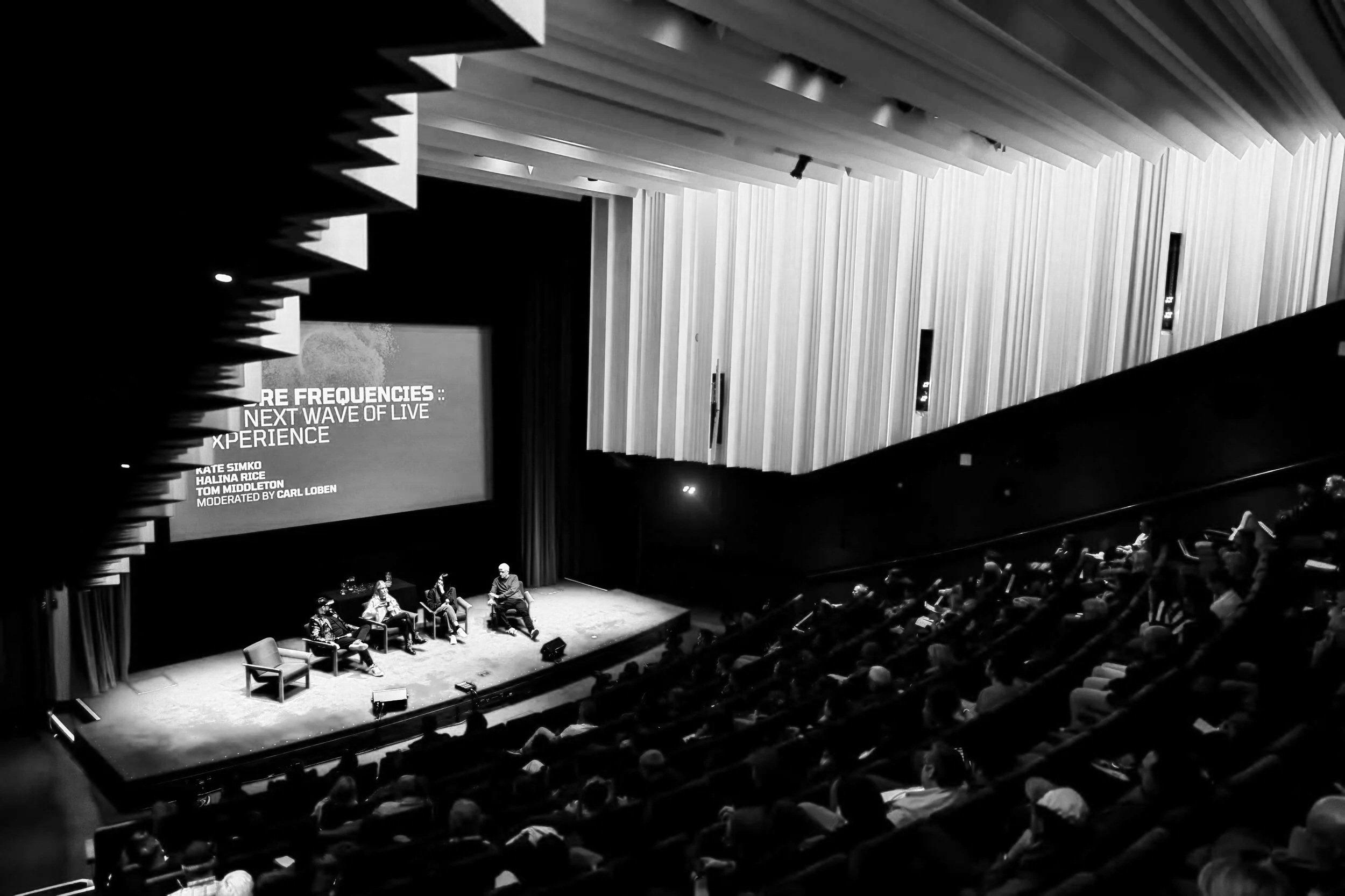 An indoor stage event with four panelists seated on stage, a large screen displaying text about music frequencies, with an audience seated in a darkened auditorium.