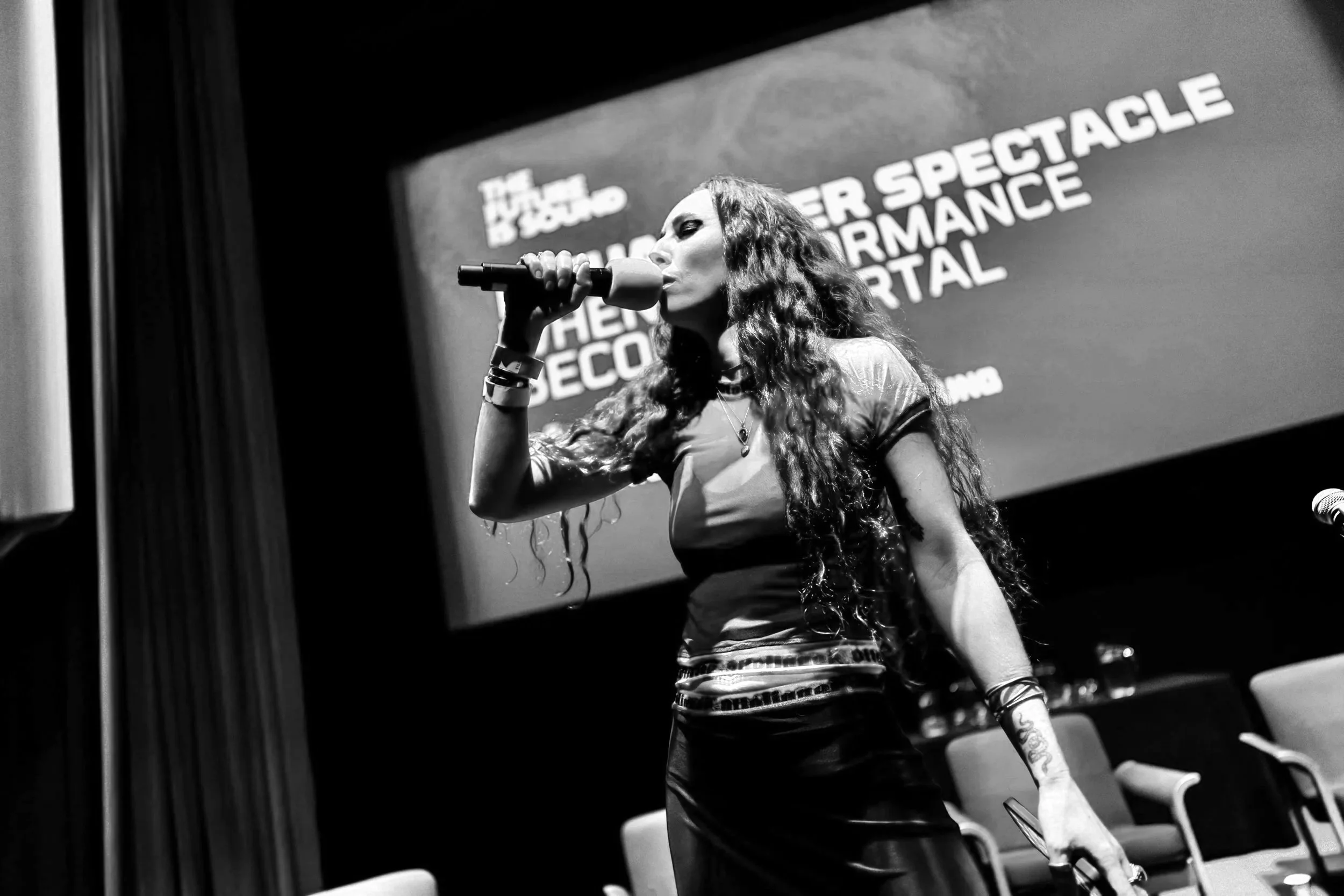 A woman with long curly red hair singing into a microphone on stage at a conference with a large screen behind her displaying the words 'The Future of the Artist' and 'Performance Portal'.