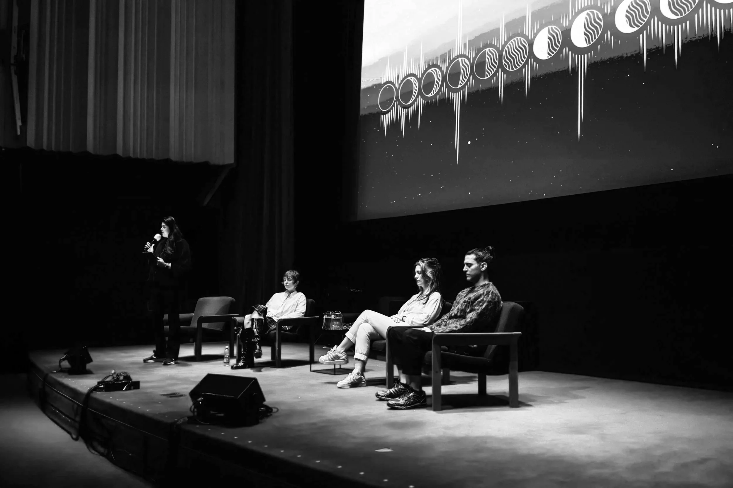 A panel discussion on a stage with four women, one standing and speaking into a microphone, and three sitting in chairs. The background features a large screen with abstract art or graphics.