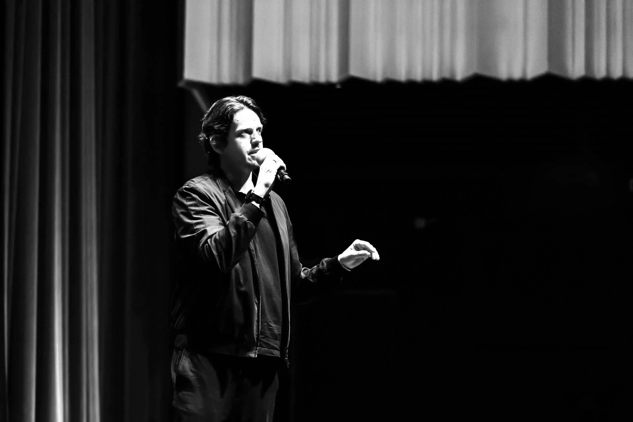 A young man with dark, wavy hair, wearing a black jacket and black shirt, speaking into a microphone on stage with red curtains in the background.