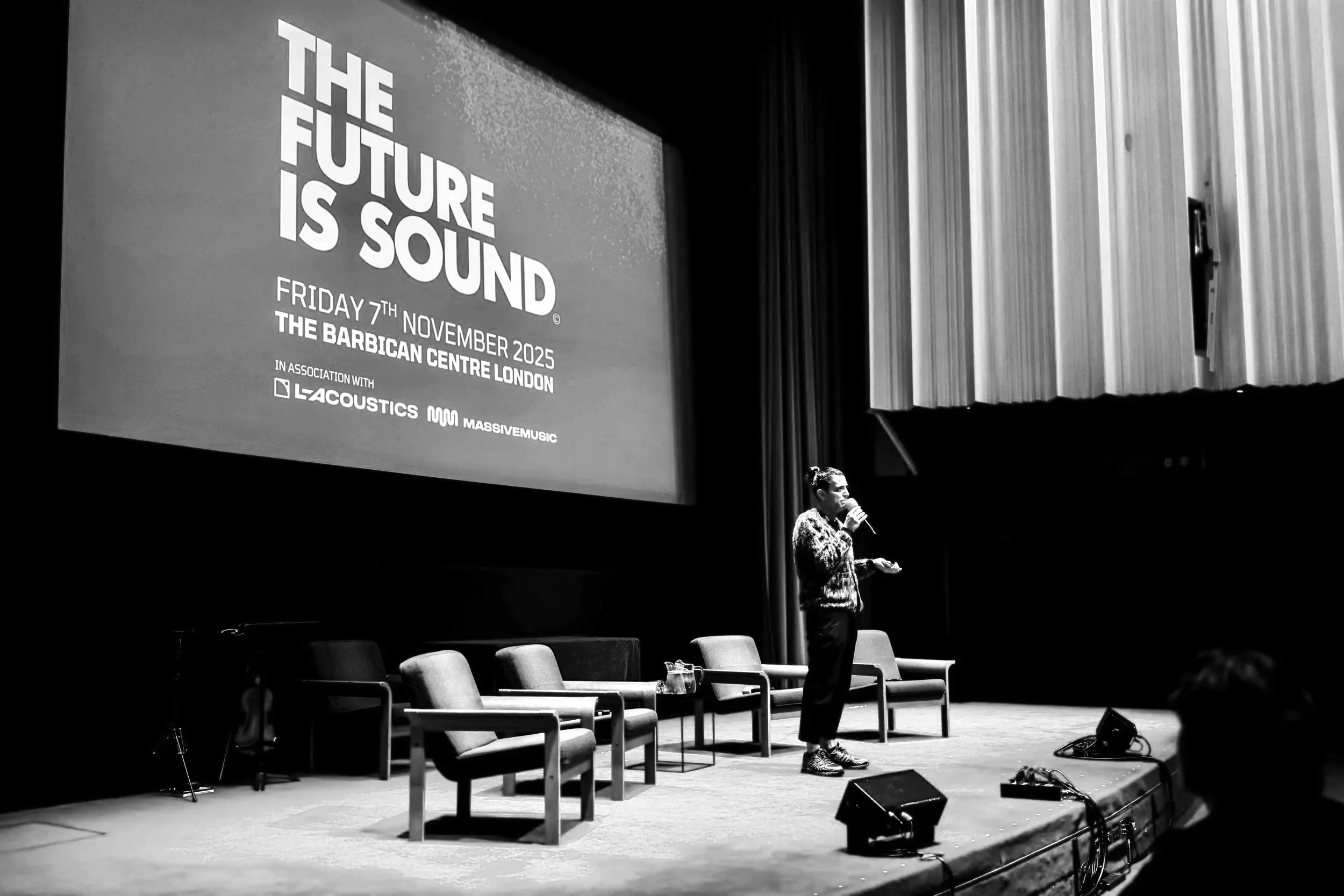 A woman on stage speaking into a microphone at an event called 'The Future is Sound' on November 7, 2025, at The Barbican Centre in London. The stage has four chairs and a small table, with a large screen displaying event details and logos of LaCoust