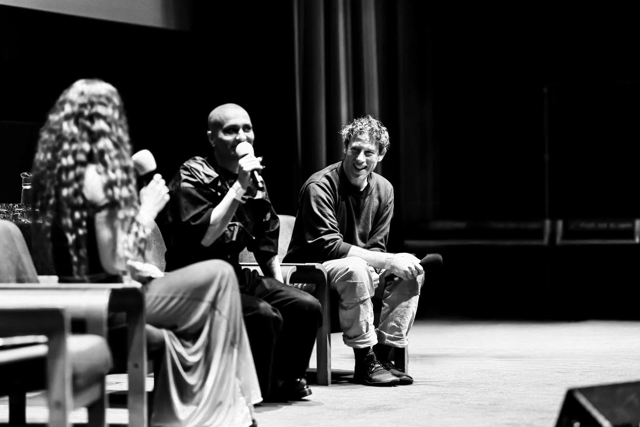 Three people sitting on chairs on stage in discussion, two women and one man, with a black background and red curtains.