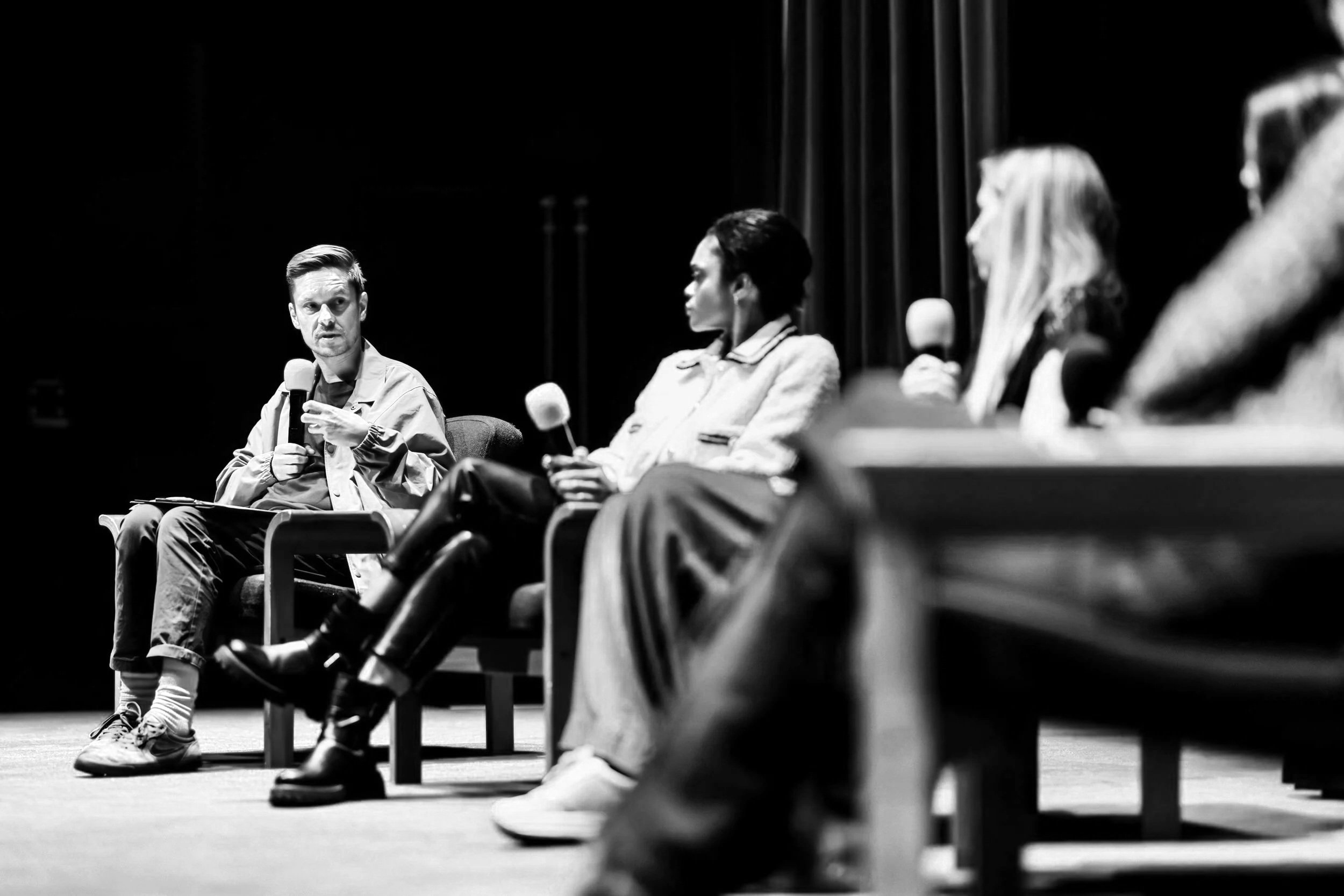 A panel discussion with four people sitting on chairs on a stage, holding microphones, with dark background and red curtains.
