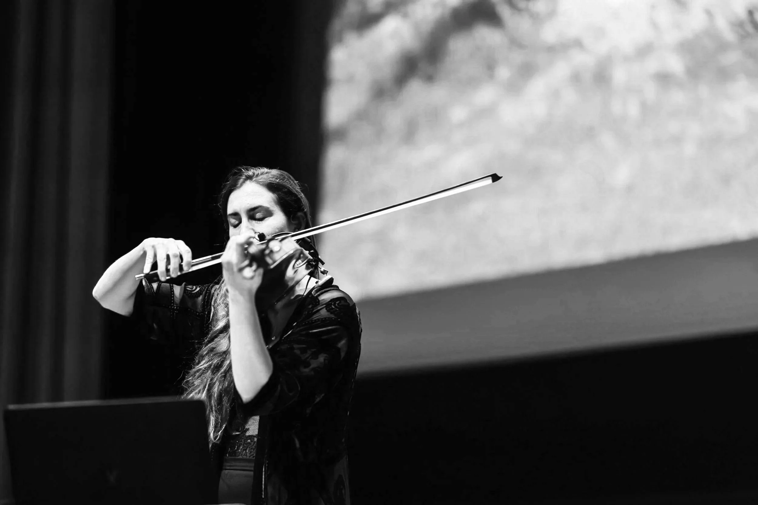 A woman playing the violin on stage, with a large screen displaying an abstract background behind her.