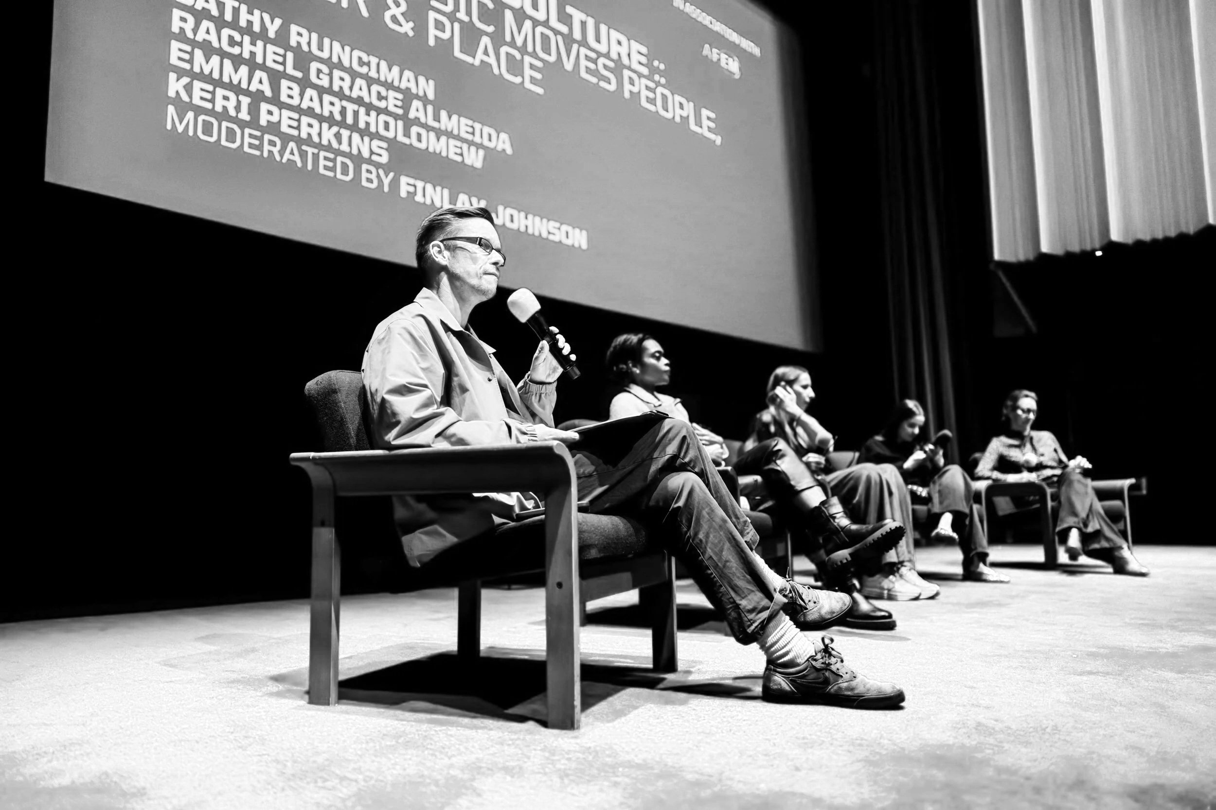 Panel discussion with five women sitting on stage in front of a large blue screen, the woman on the far left holding a microphone with an orange foam cover, all dressed casually, on a red carpeted stage with red curtains on the right.