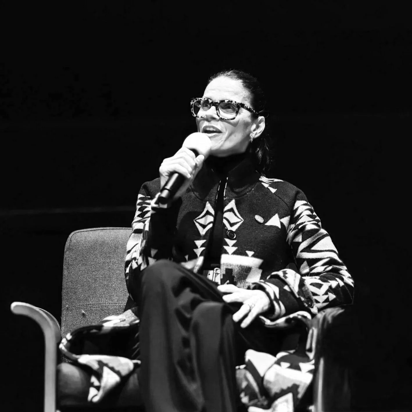 A woman wearing glasses and a patterned jacket is sitting on a chair, holding a microphone.
