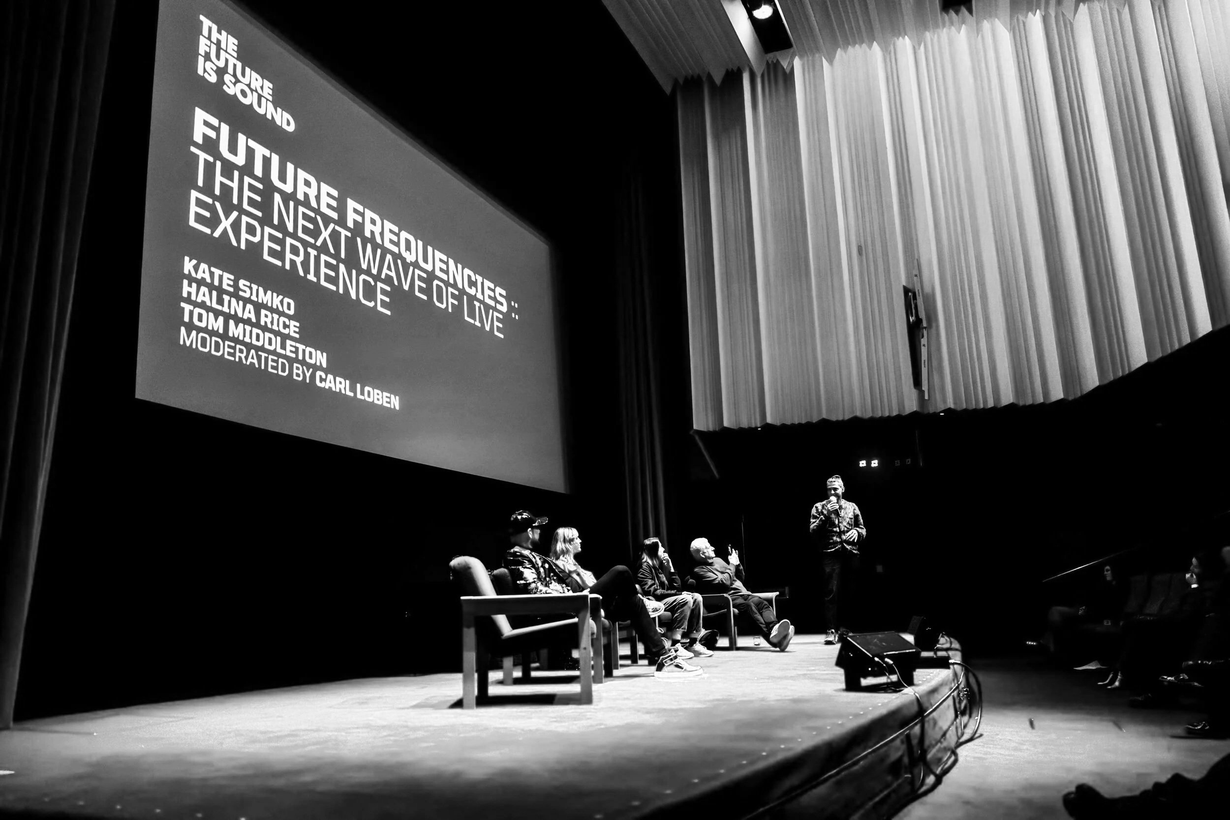 Panel discussion at a conference with five speakers sitting on chairs and one standing on stage, a large screen displaying the event title and moderator's name, red carpeted stage, and audience members visible.